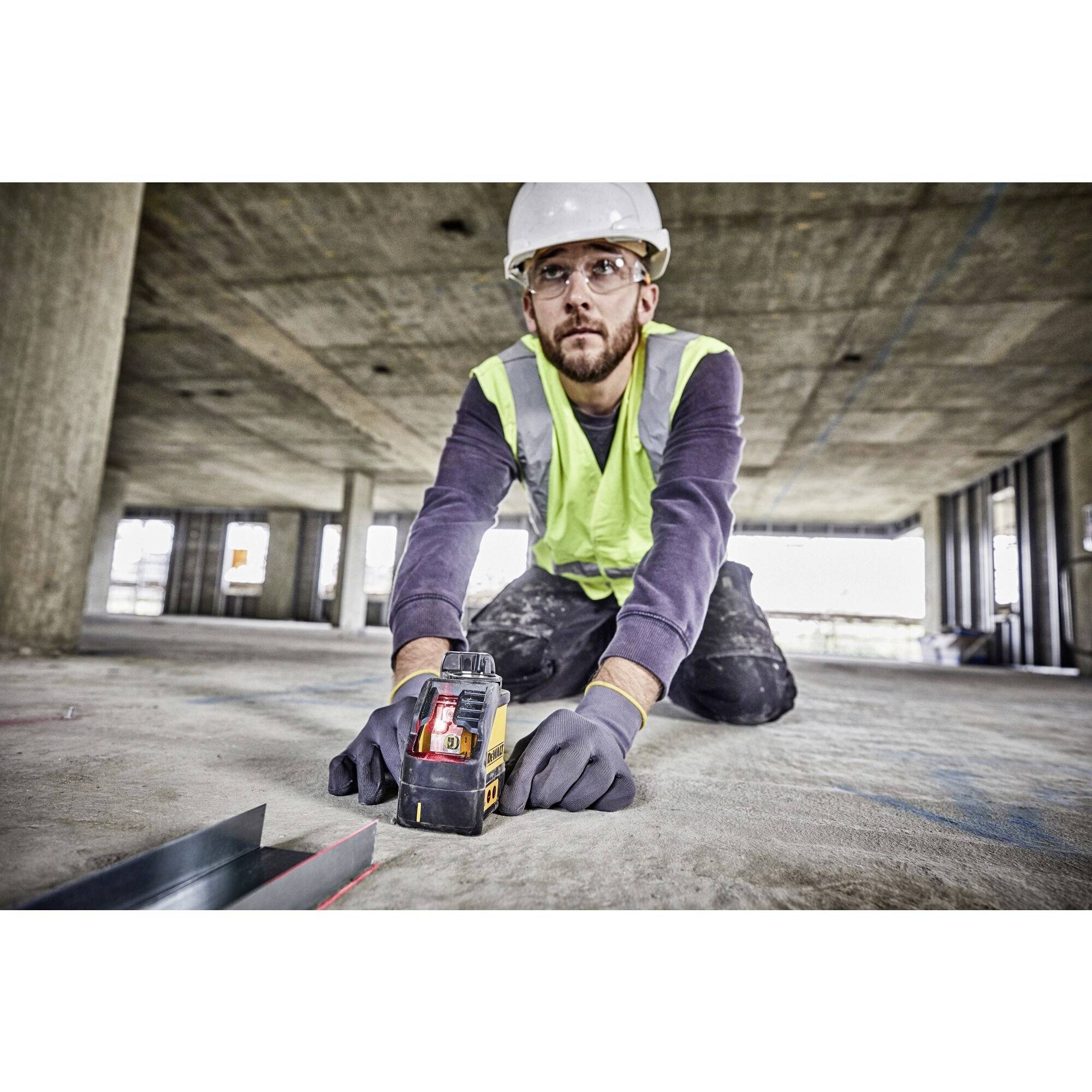 Arbeiter auf Baustelle im Rohbau, kniend und fokussiert, trägt Schutzkleidung inklusive Weste, Helm und Handschuhe, nutzt Werkzeug am Boden.