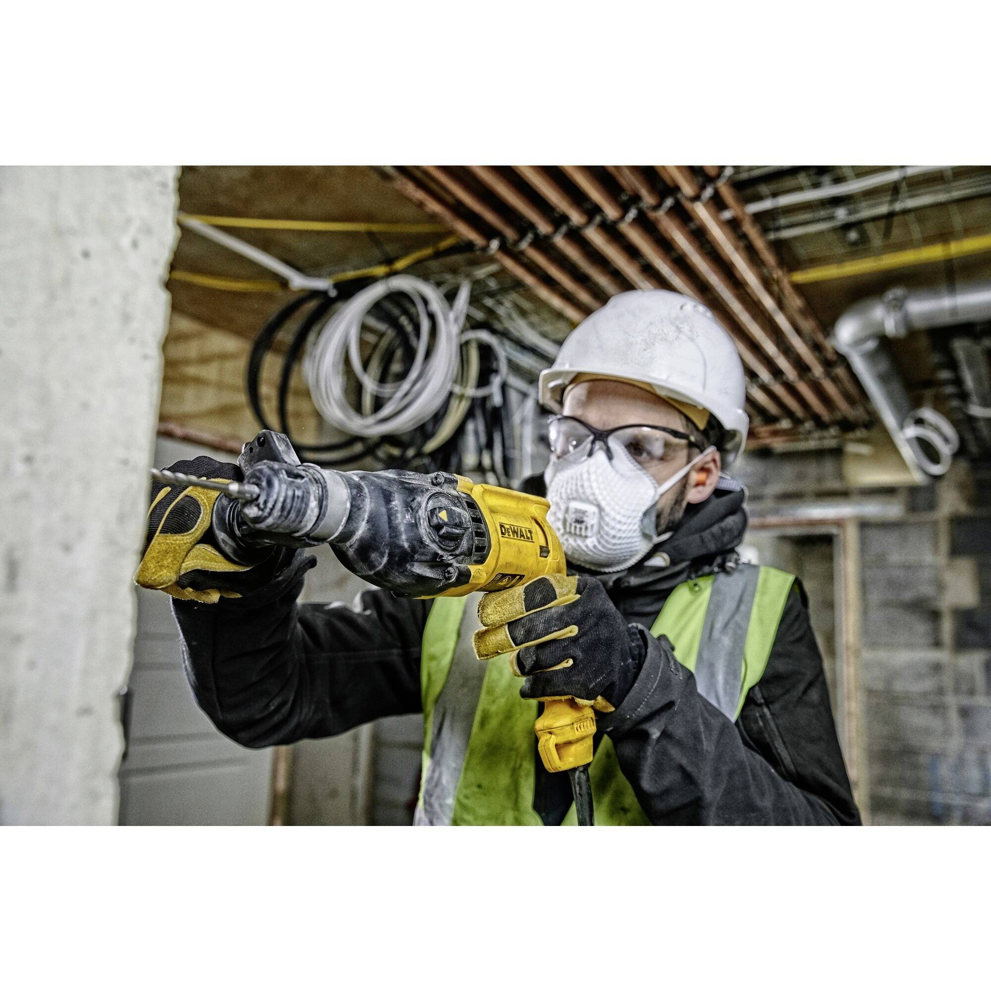 Ein Bauarbeiter mit Schutzmaske, Helm und Warnweste verwendet eine elektrische Bohrmaschine auf einer Baustelle mit sichtbaren Rohren im Hintergrund.