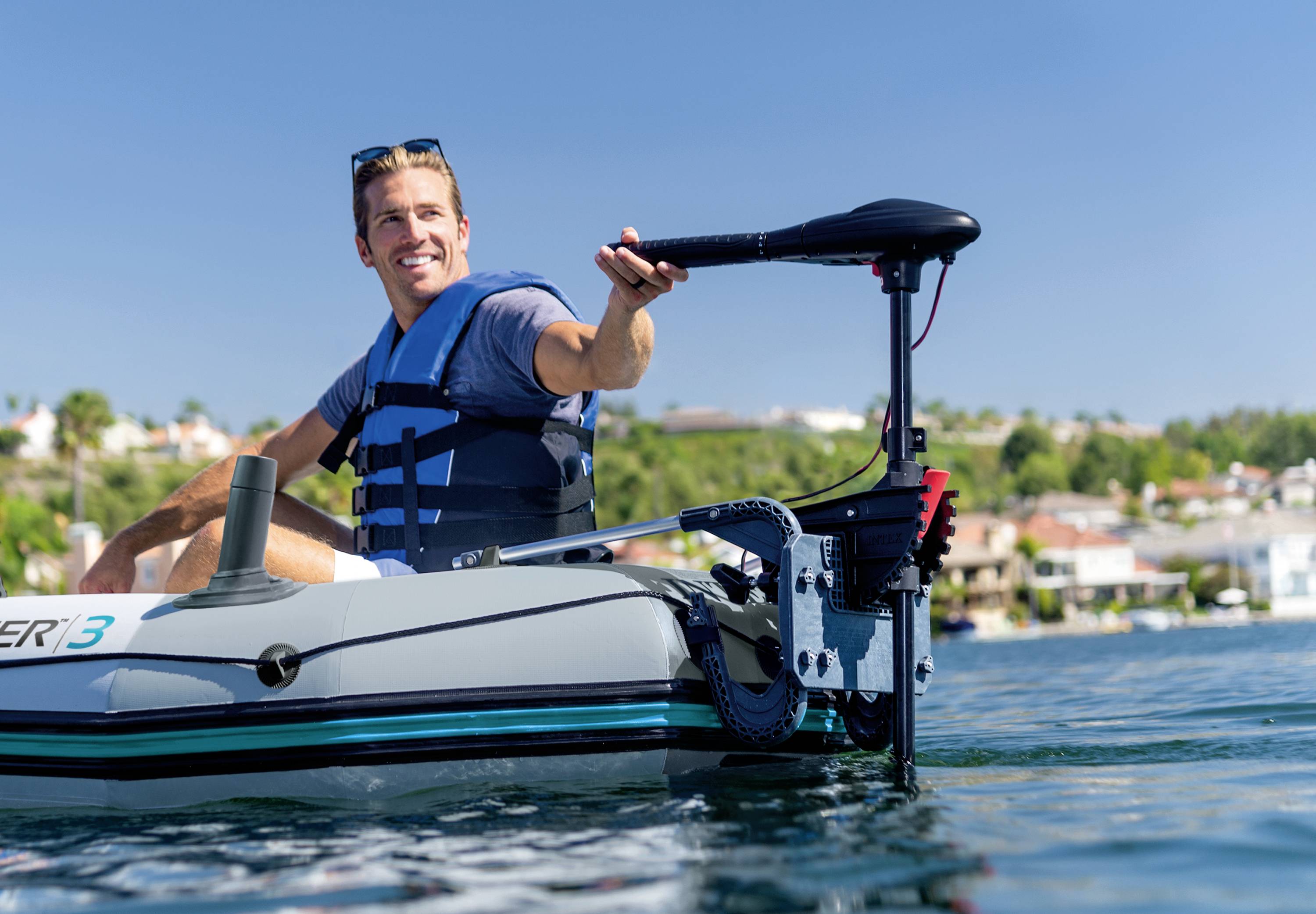 Person in einem aufblasbaren Boot auf einem See, trägt eine Schwimmweste und hält den Steuergriff eines Elektromotors. Im Hintergrund Wohnhäuser.