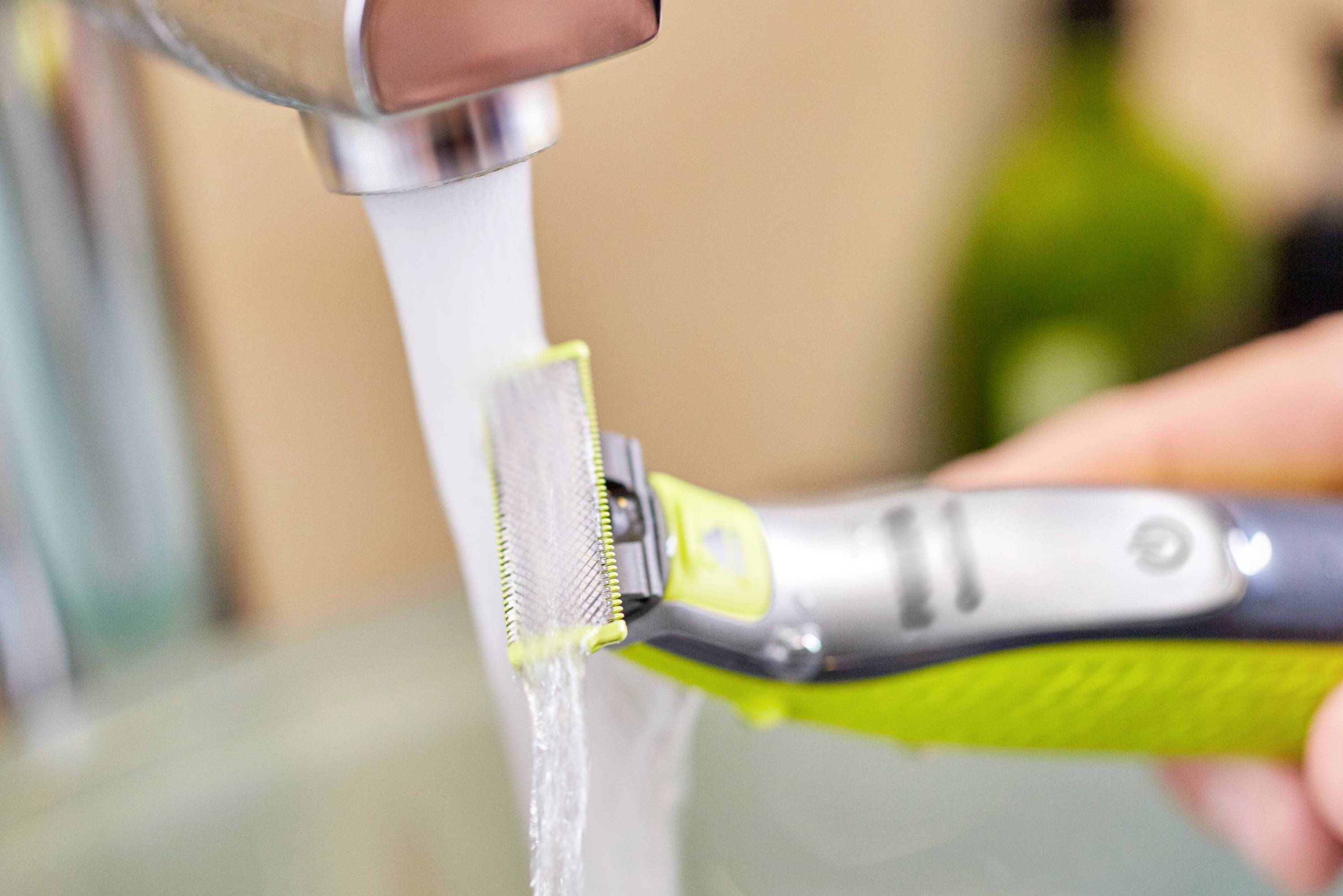 Elektrischer Rasierer wird unter fließendem Wasser gereinigt. Hand hält Rasierer unter Wasserhahn, Hintergrund unscharf.