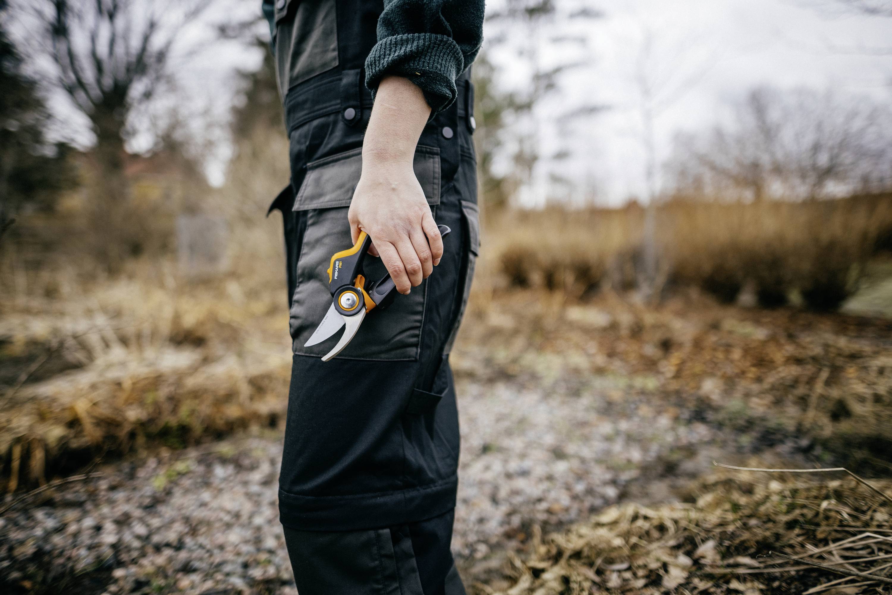 Eine Person hält eine Gartenschere in der Hand, während sie in einem herbstlichen Garten steht. Die Szene vermittelt Gartenarbeit im Herbst.