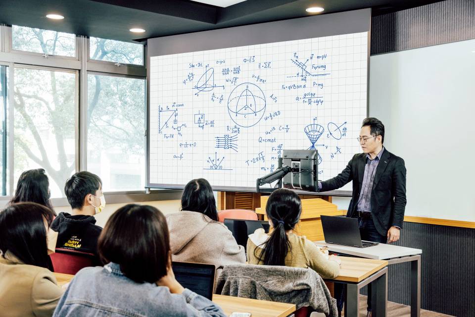 Ein Lehrer erklärt einer Gruppe Studierender an einem Whiteboard mathematische Formeln und Diagramme in einem Klassenraum.