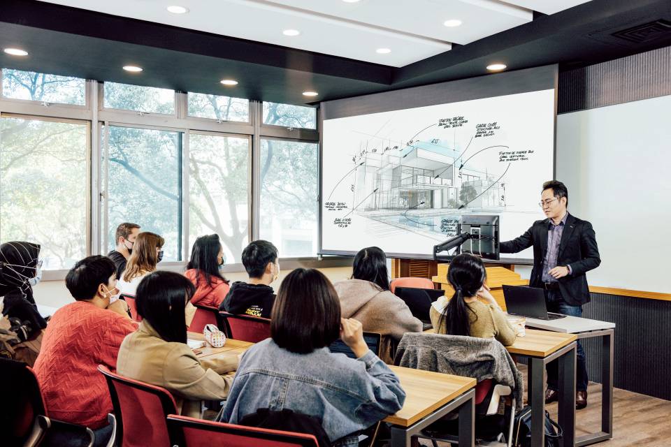 Dozent präsentiert Architekturentwurf auf Leinwand vor Studierenden in einem Klassenzimmer. Die Studierenden hören aufmerksam zu.