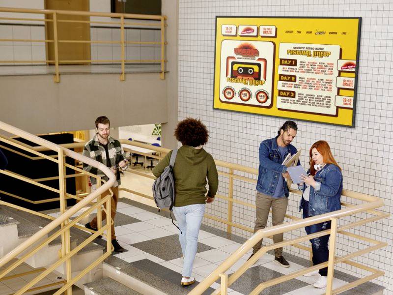 Vier Personen in einem Treppenhaus, zwei schauen auf ein Tablet und diskutieren. An der Wand hängt ein großes, buntes Poster.