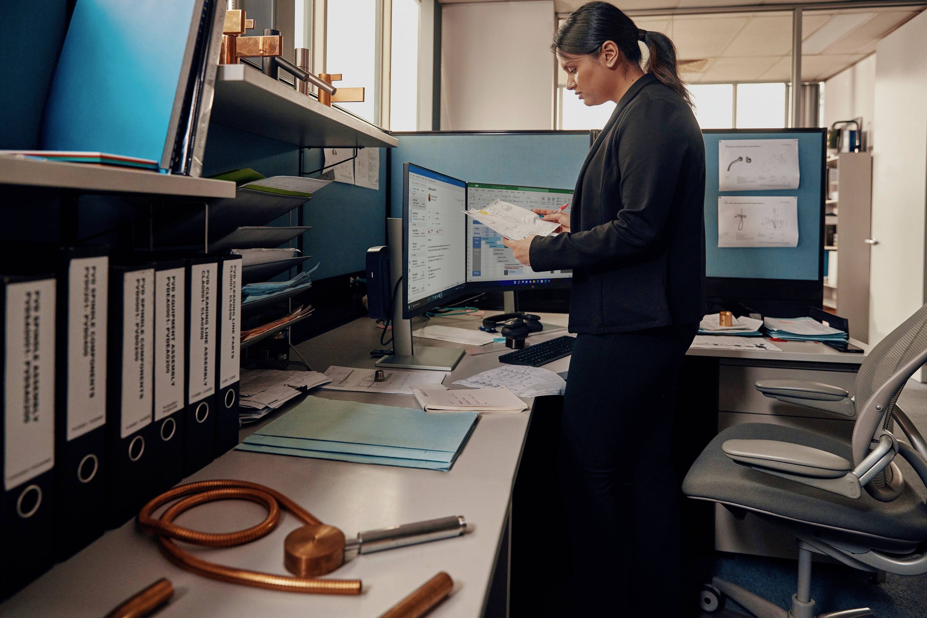 Eine Person in einem Büro prüft Dokumente vor zwei Computerbildschirmen. Im Vordergrund liegen Büromaterialien und Baupläne.
