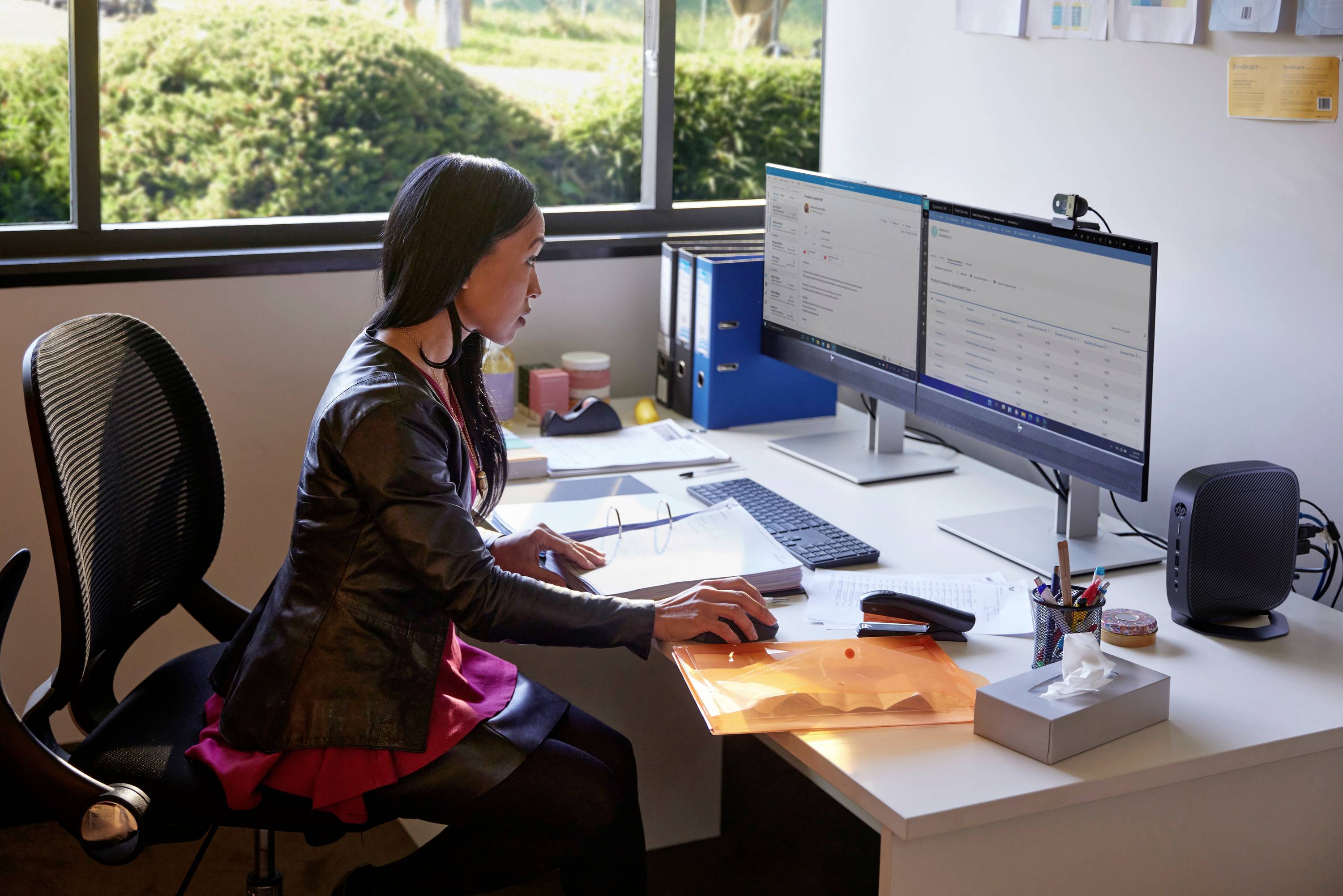 Eine Person sitzt an einem Schreibtisch mit zwei Monitoren und arbeitet am Computer. Auf dem Tisch befinden sich Ordner und Bürobedarf.