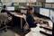 Zwei Personen arbeiten in einem Büro an Computern. Beide sitzen an Schreibtischen mit Monitoren, Ordnern und Papieren.