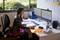 Eine Frau sitzt an einem Schreibtisch und arbeitet am Computer. Sie trägt eine schwarze Jacke und ein pinkfarbenes Kleid. Im Hintergrund sind Pflanzen und Ordner zu sehen.