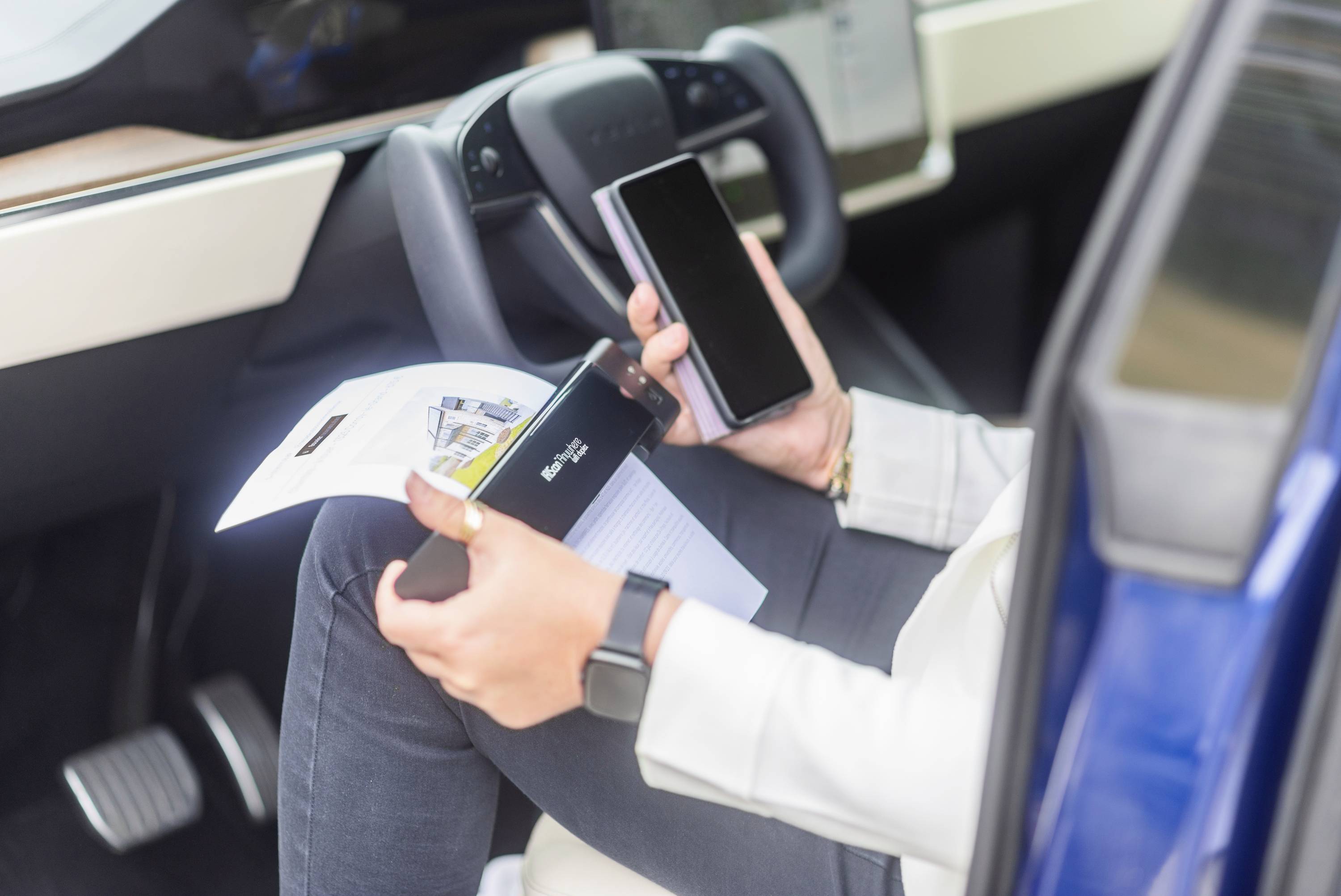 Person im Auto, hält Smartphone und Dokument. Lenkrad und Armaturenbrett im Hintergrund sichtbar. Moderne Technologie und Mobilität.