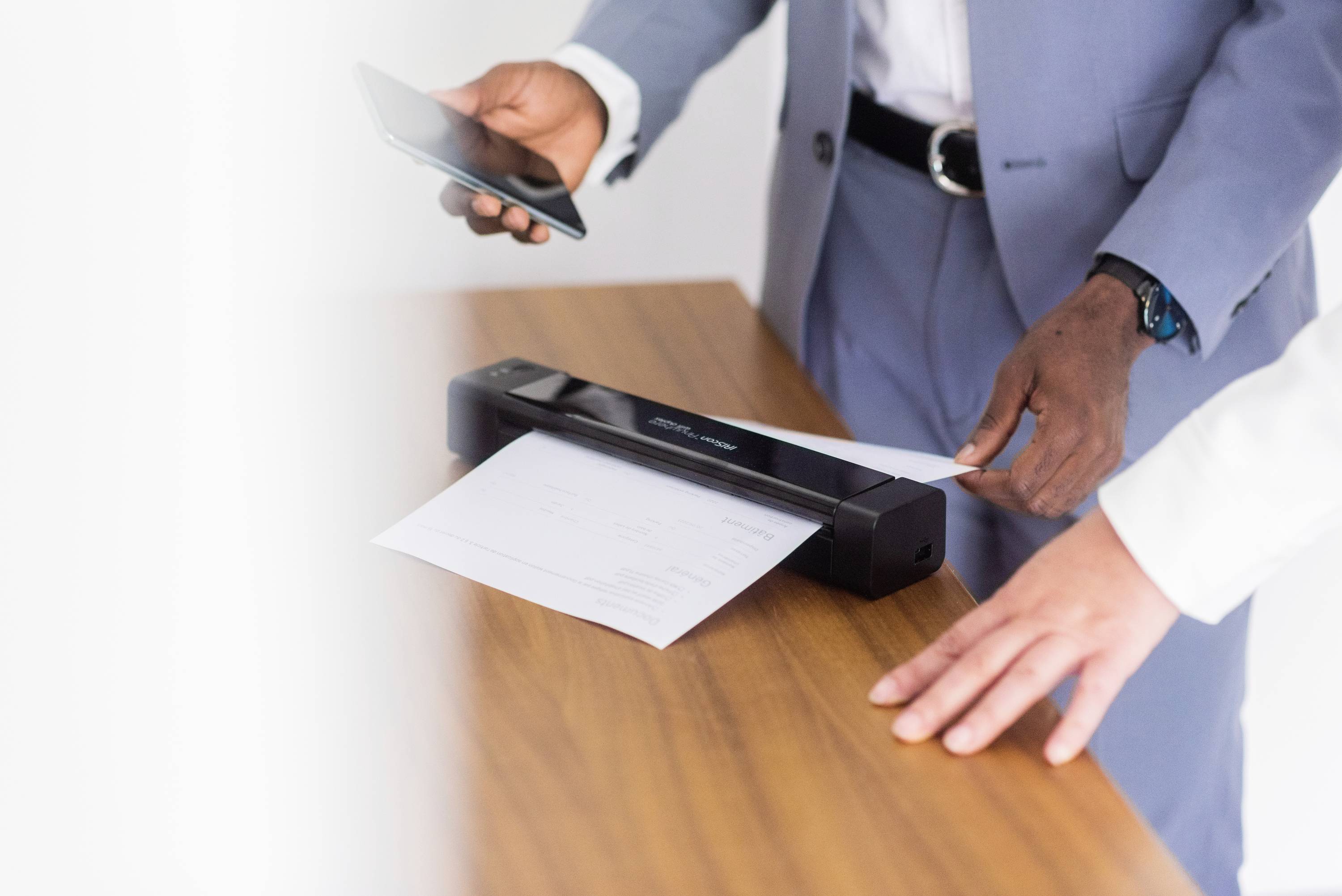 Zwei Personen scannen ein Dokument mit einem mobilen Scanner auf einem Holztisch. Eine Person hält ein Smartphone in der Hand.