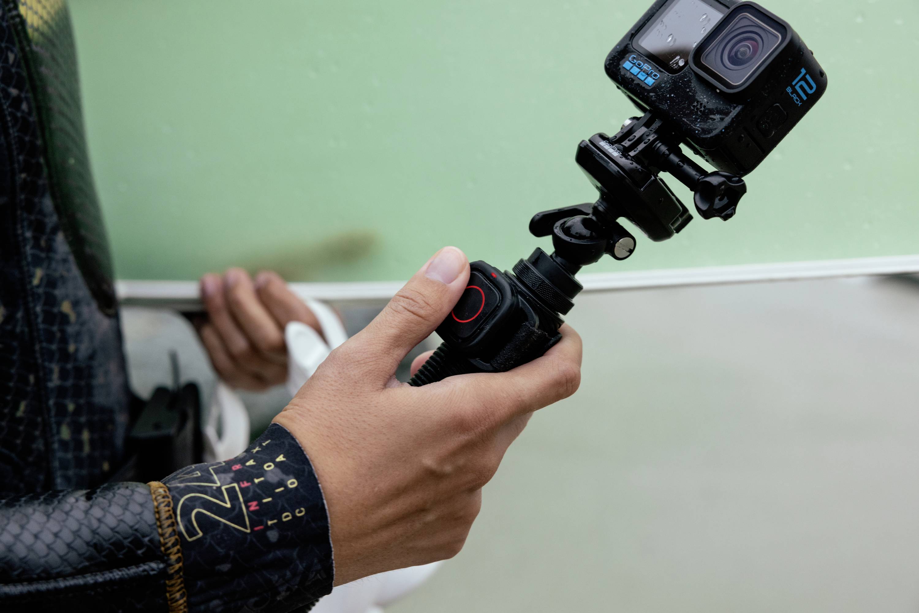 Eine Person hält eine Action-Kamera mit Halterung, bereit zum Filmen. Im Hintergrund ist unscharf ein Surfbrett zu erkennen.