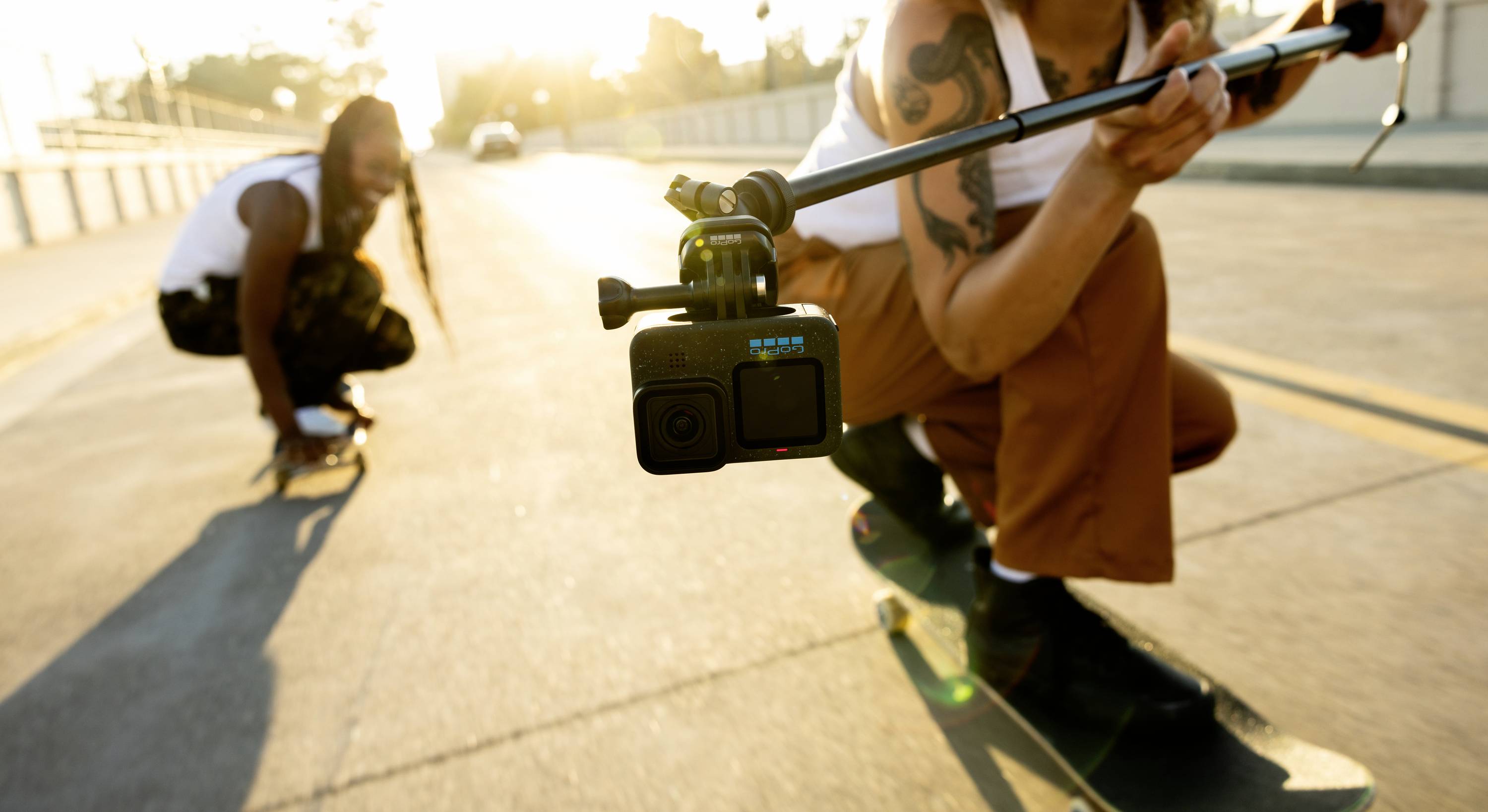 Zwei Personen skaten auf einer Straße bei Sonnenuntergang, während sie sich mit einer Kamera auf einem Selfie-Stick filmen. Stimmung dynamisch und lebendig.