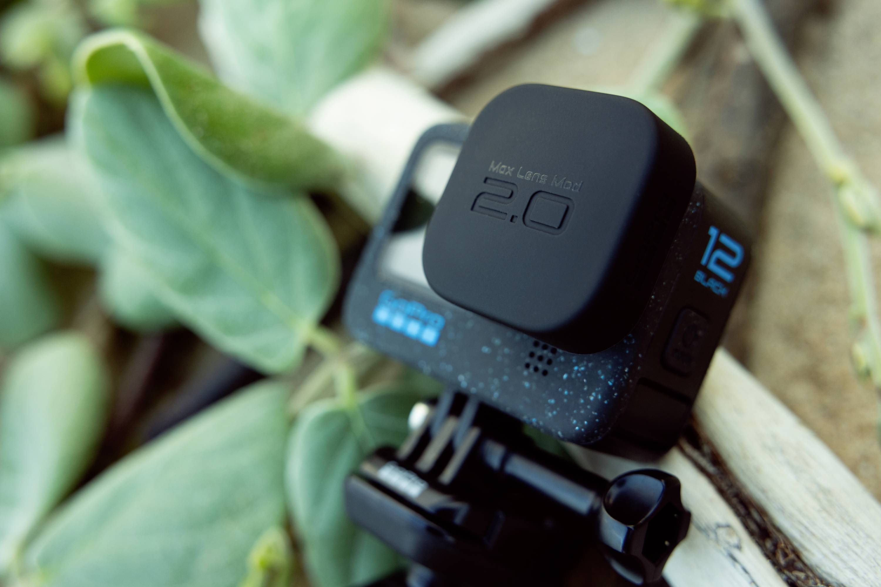 Eine Action-Kamera mit Verpackung 'Max Lens Mod 2.0' liegt auf einem weißen Ast neben grünen Blättern.