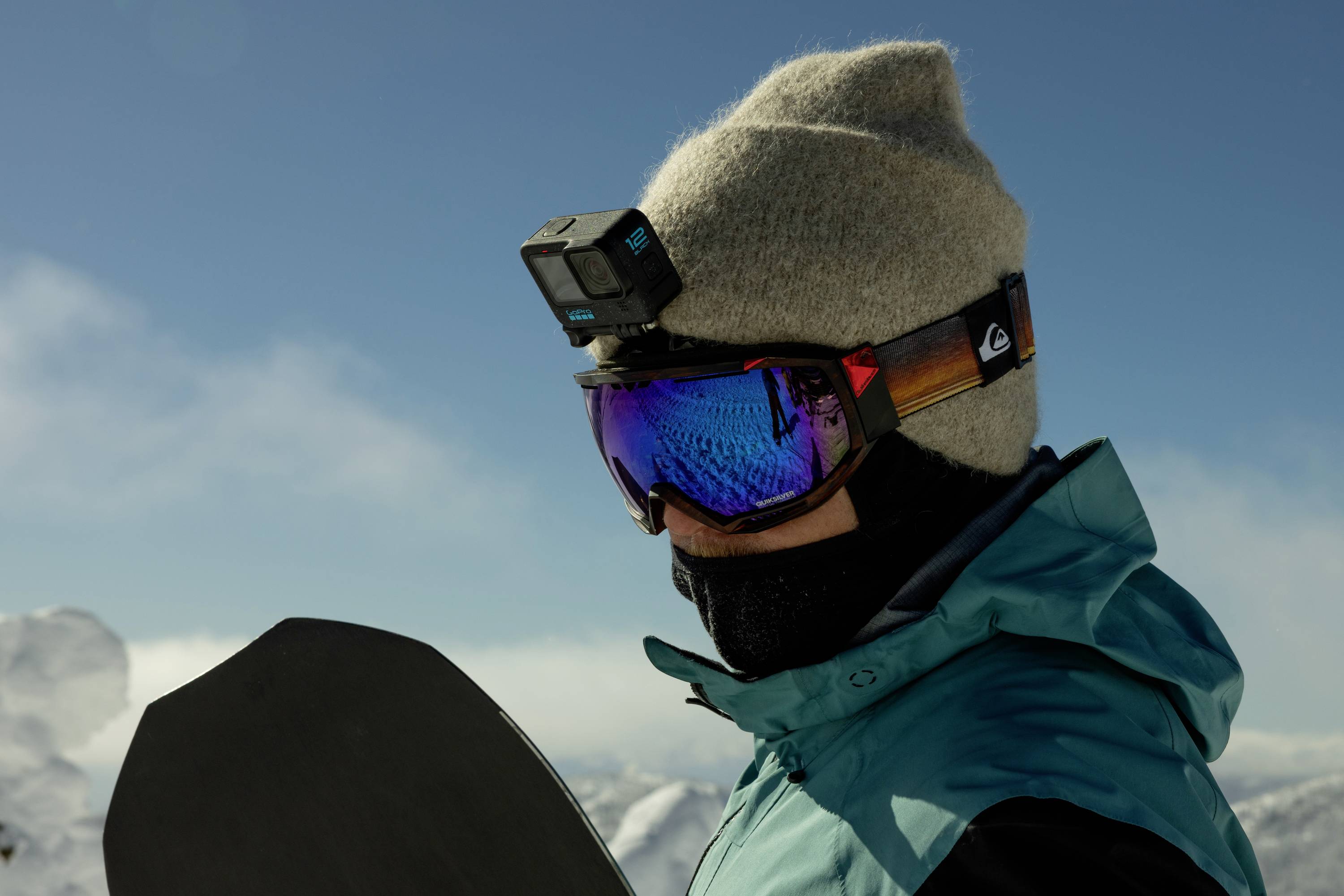 Eine Person in Winterkleidung mit Skibrille und Kamera auf dem Helm schaut auf ein Snowboard in schneebedeckter Berglandschaft.