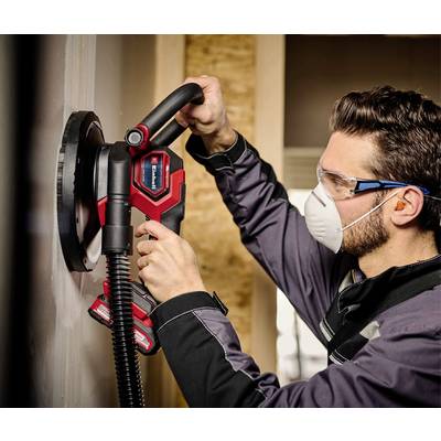 Ein Mann trägt eine Schutzbrille und Maske und schleift mit einer elektrischen Schleifmaschine eine Wand.