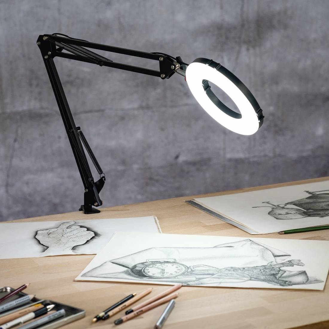 Zeichentisch mit Zeichnungen, einem flexiblen Leuchtring darüber; Bleistifte und Radiergummi im Vordergrund.
