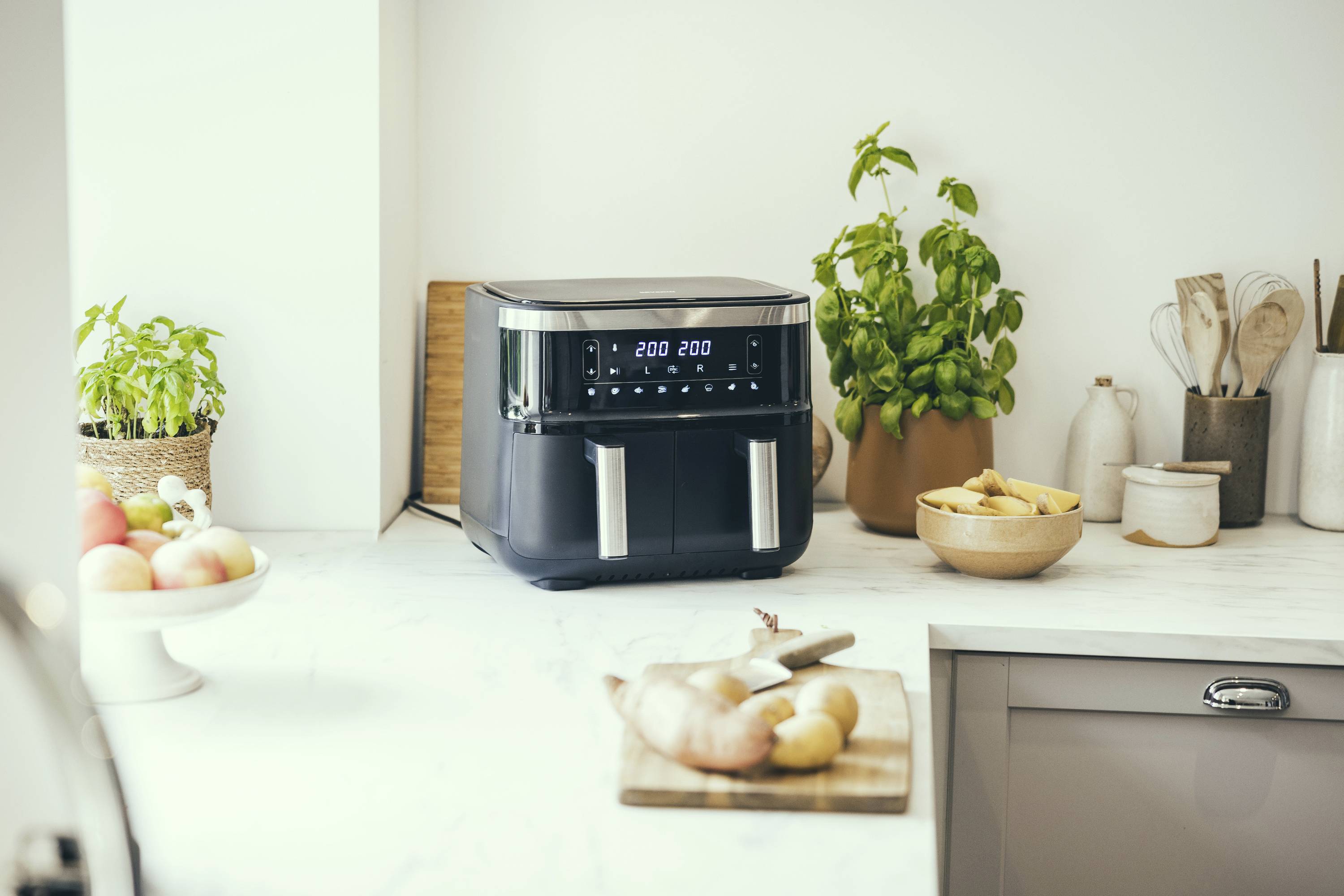 Digitale Heißluftfritteuse auf der Küchentheke mit Obst, Gemüsebrett und Topfpflanzen im Hintergrund.