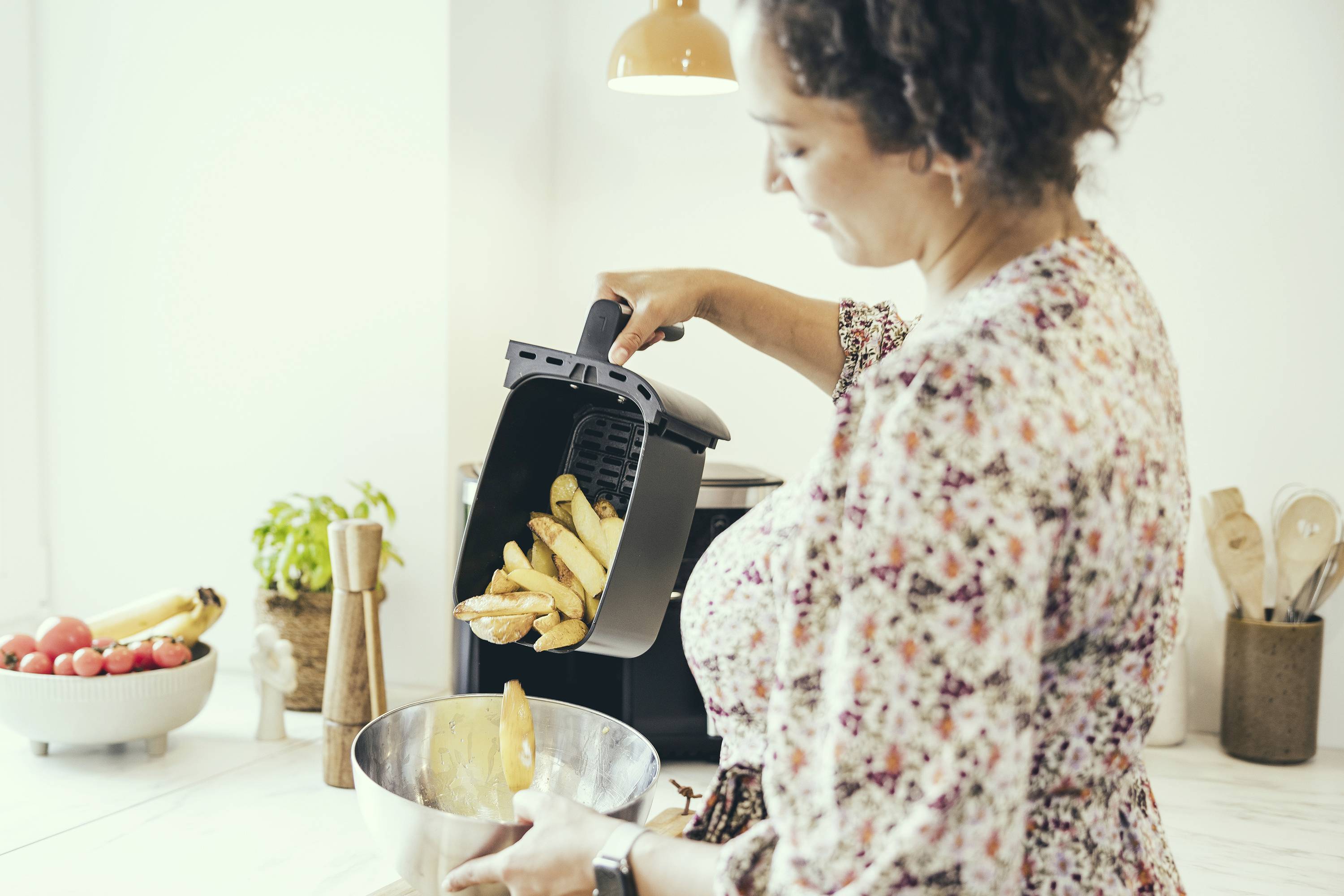 Frau kippt frisch gebackene Kartoffelspalten aus einer Heißluftfritteuse in eine Schüssel. Im Hintergrund ist eine Küche zu sehen.