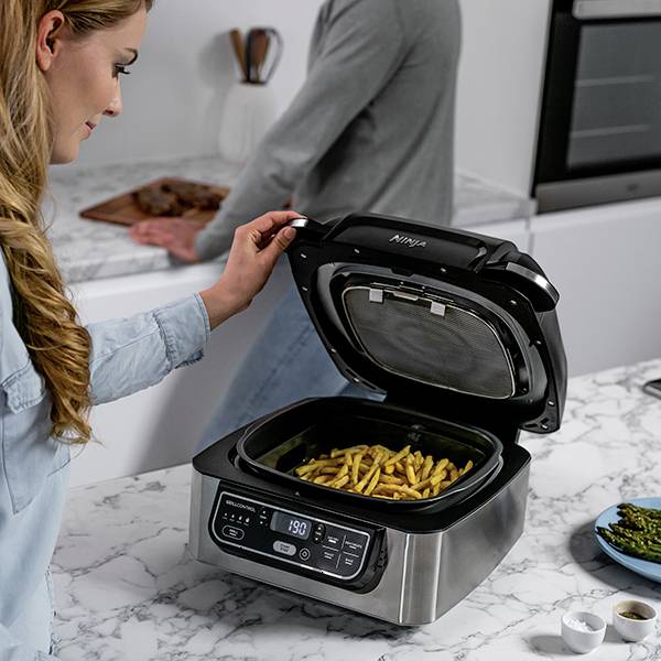 Eine Frau öffnet eine Heißluftfritteuse mit Pommes frites auf einer Küchenzeile, während eine Person im Hintergrund kocht.