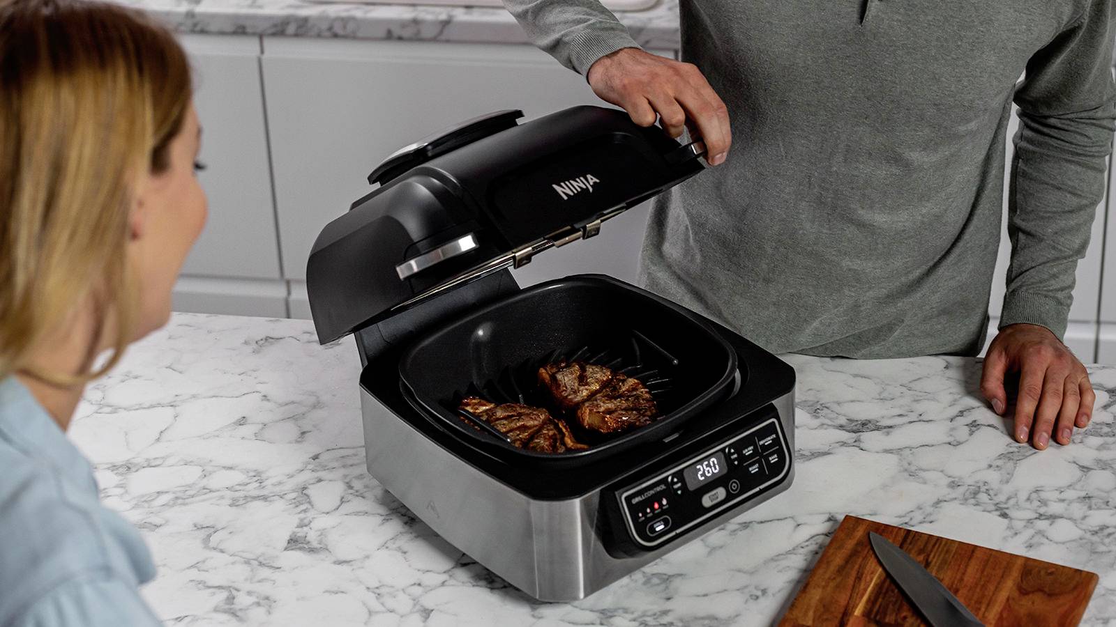 Eine Person grillt Fleischstücke auf einem Tischgrill in einer Küche. Eine andere Person schaut zu. Der Grill zeigt 200 °C an.