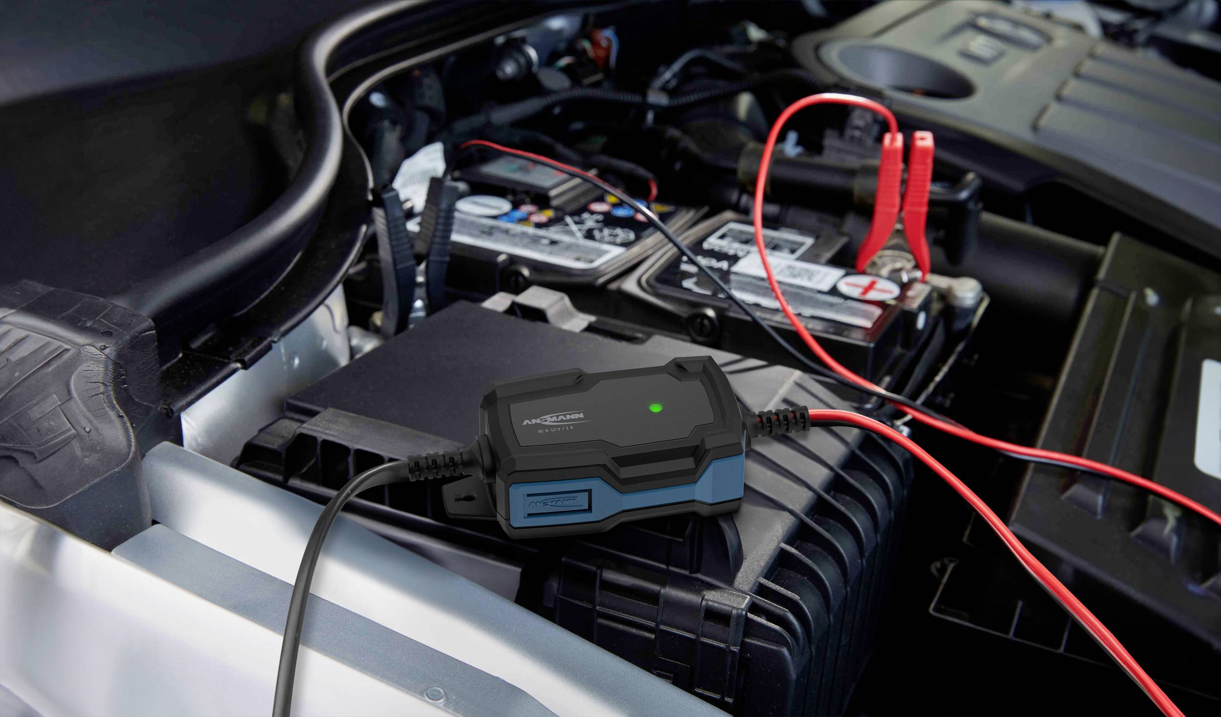 Ein Batterieladegerät ist an eine Autobatterie im Motorraum angeschlossen, mit roten und schwarzen Kabeln, die die Verbindung zeigen.