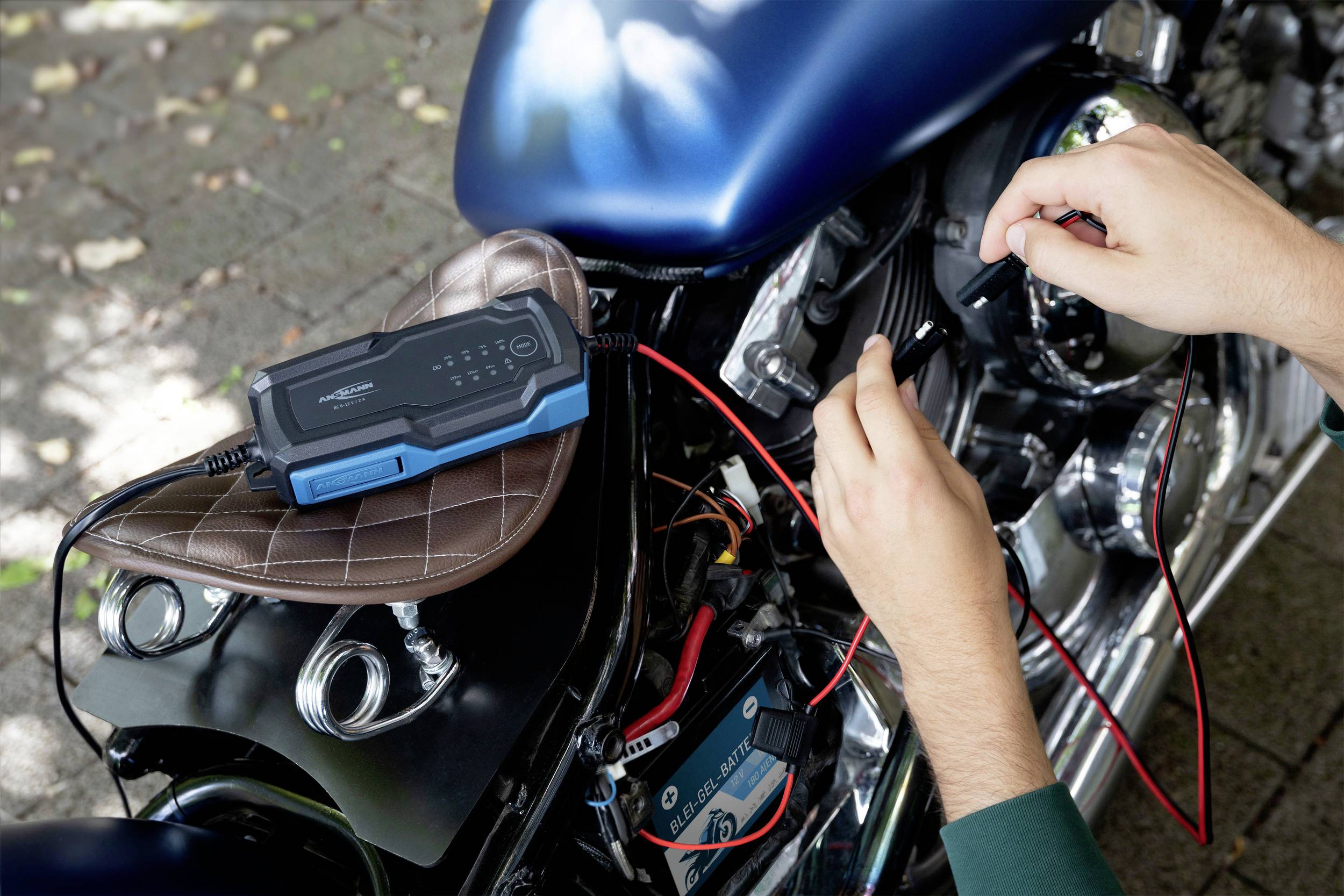 'Ein Ladegerät ist mit einem Motorrad verbunden. Zwei Hände halten die Kabel an den Batterieklemmen. Der Sitz ist braun.'