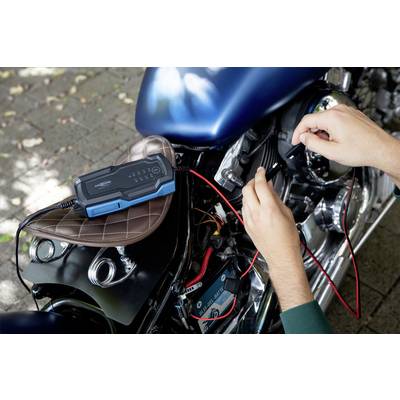 'Ein Ladegerät ist mit einem Motorrad verbunden. Zwei Hände halten die Kabel an den Batterieklemmen. Der Sitz ist braun.'