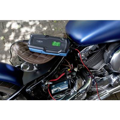 Motorrad steht auf einem Gehweg, verbunden mit einem Batterieladegerät. Das Display zeigt '12.5V' an. Schatten von Bäumen im Hintergrund.