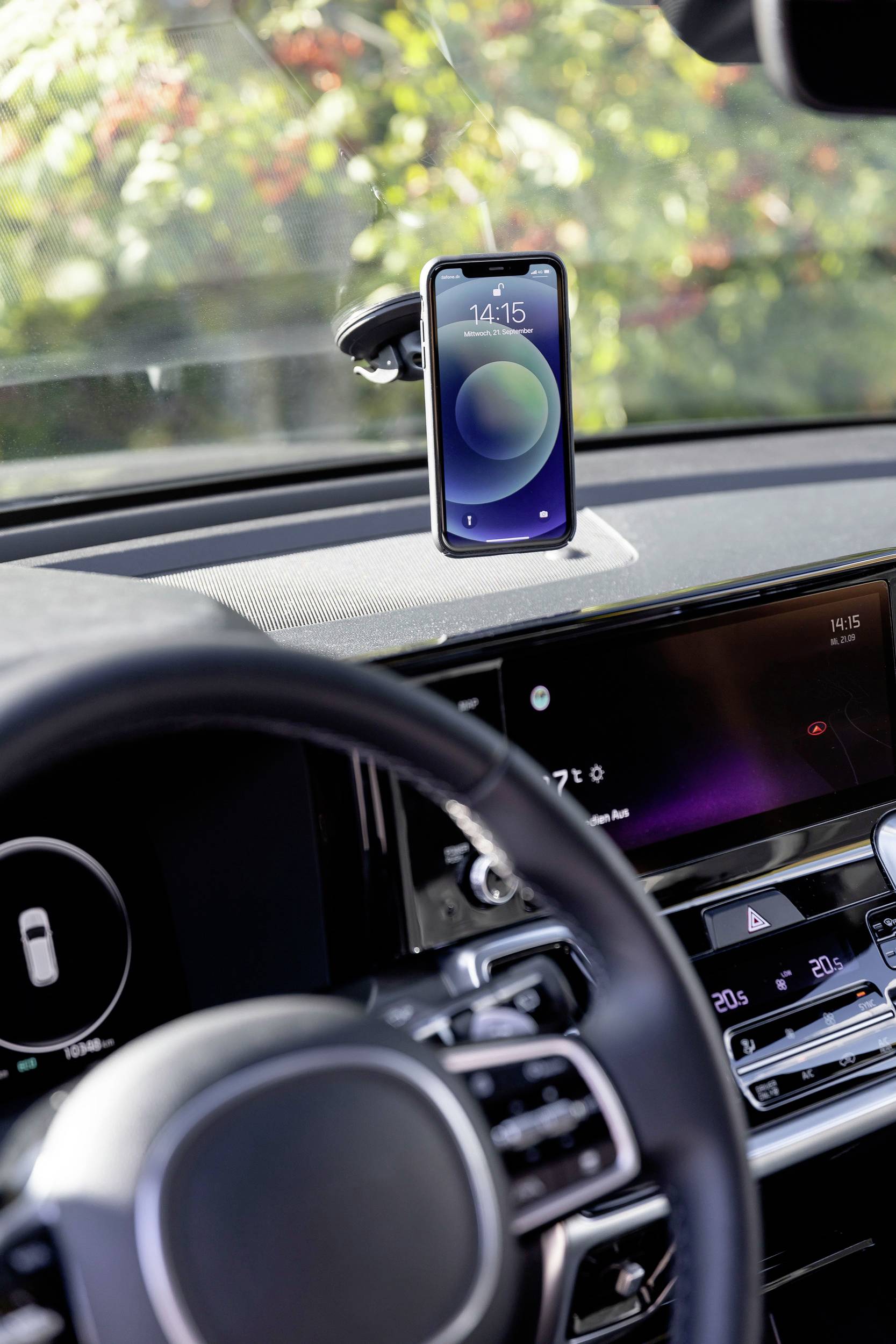 Smartphone auf einer Halterung im Auto, zeigt eine Uhrzeit an. Lenkrad und ein Teil des Armaturenbretts im Vordergrund sichtbar. Hintergrund verschwommen.