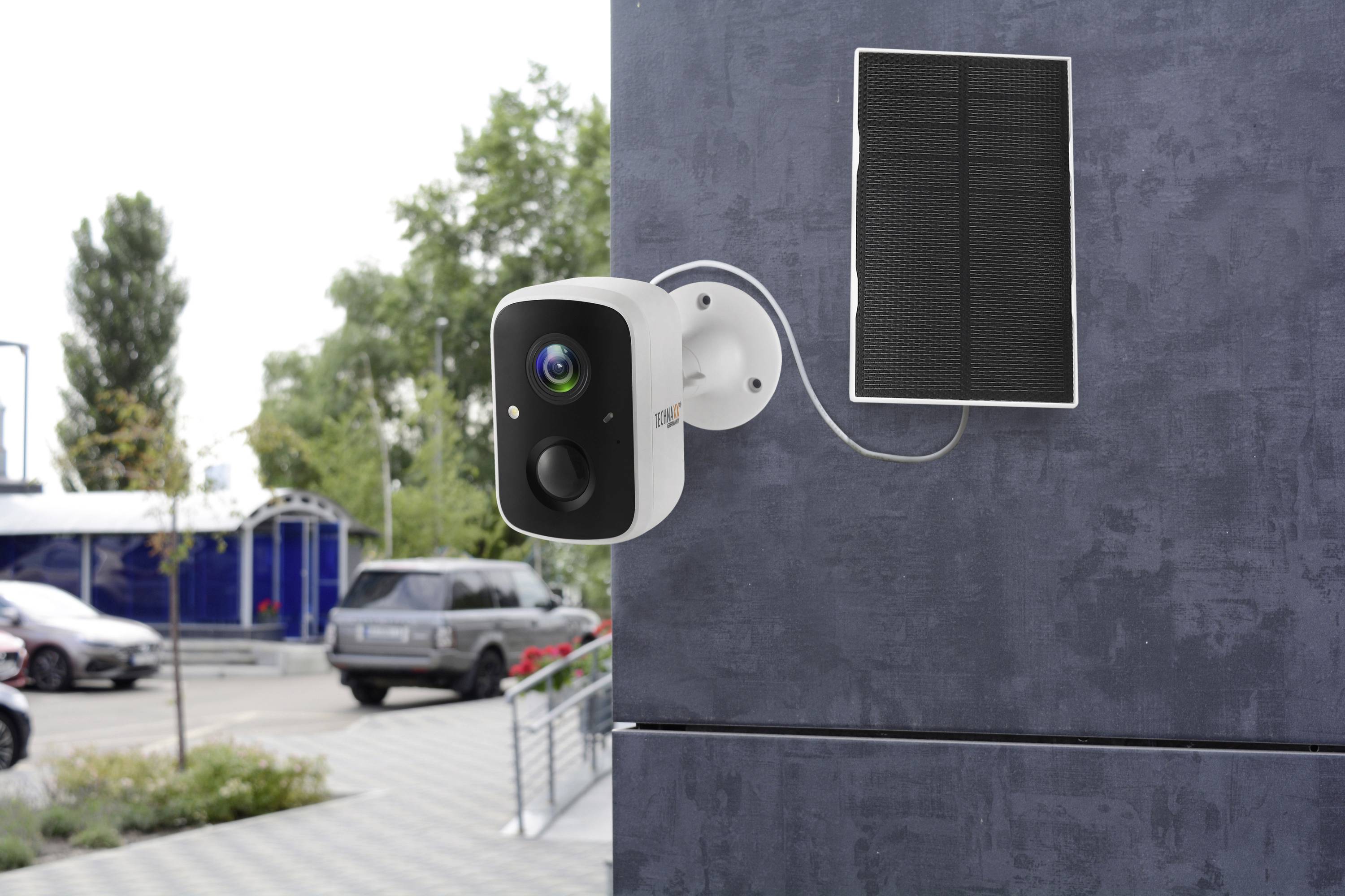 Überwachungskamera an einer Wand befestigt, mit Solarmodul verbunden. Autos und Gebäude im Hintergrund deuten auf einen Parkplatz hin.