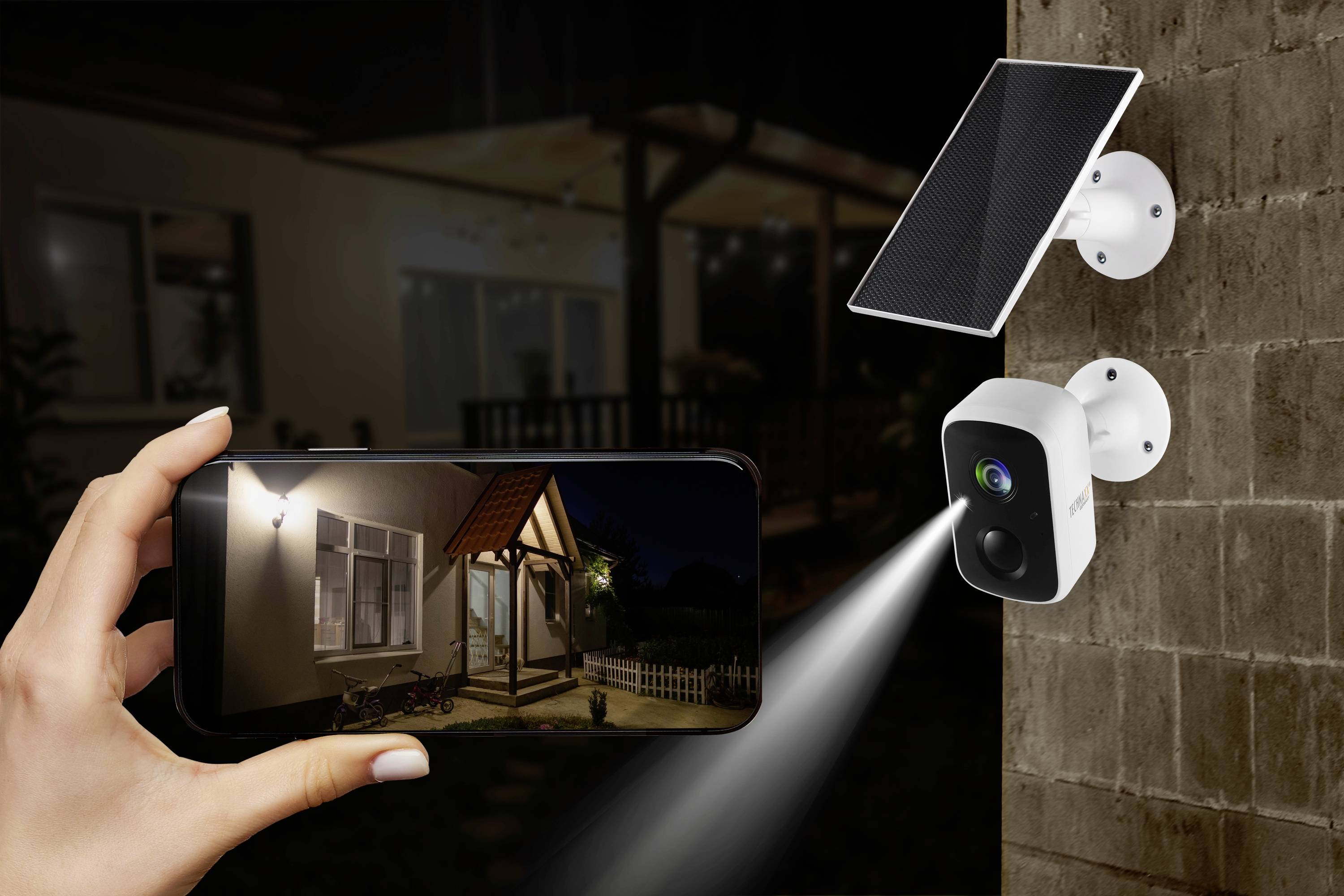 Überwachungskamera mit Solarpanel strahlt Licht auf eine Wand. Eine Hand hält ein Smartphone mit einem Überwachungsbild eines Hauses.