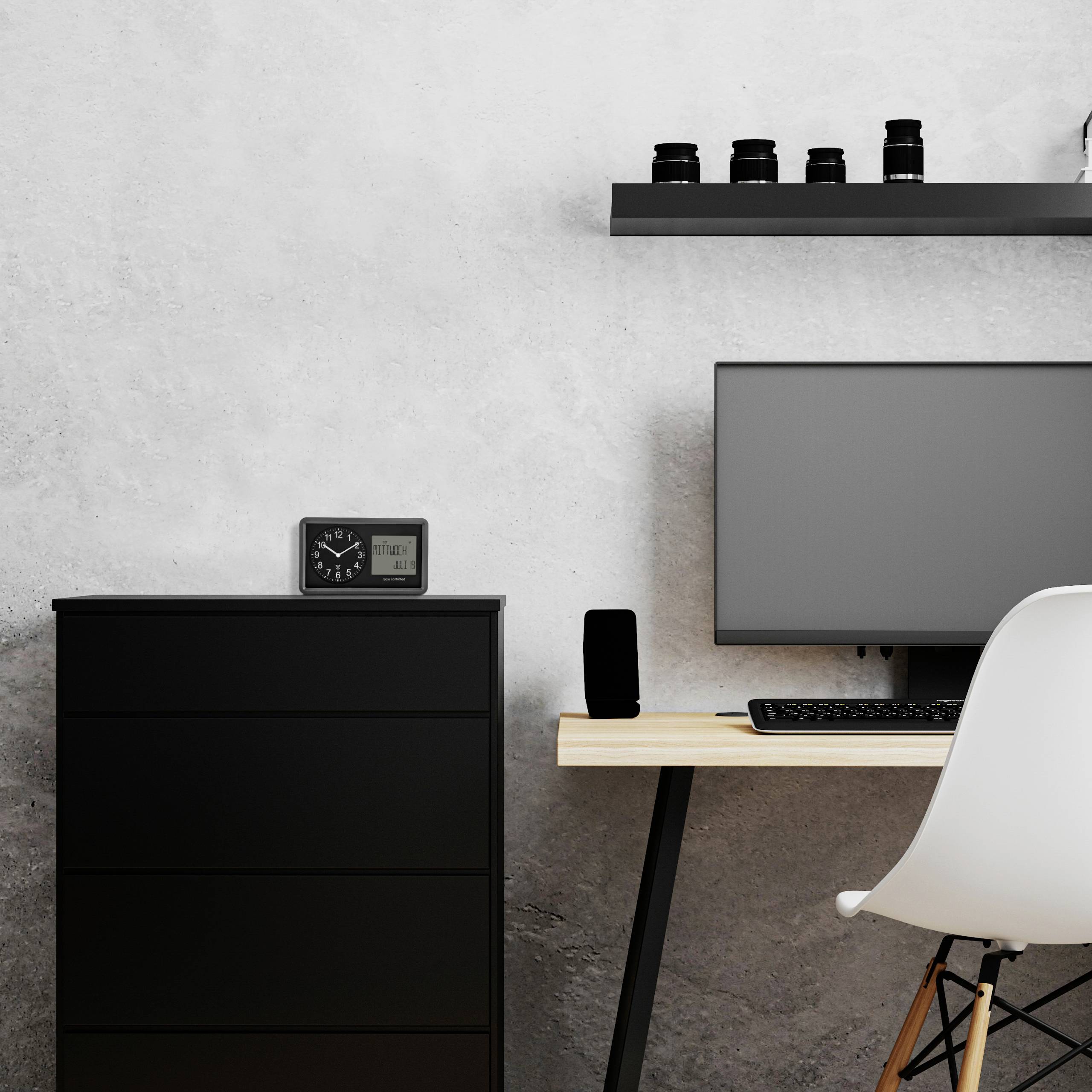 Arbeitsplatz mit Computer auf Tisch, schwarzem Stuhl, Uhr auf Schrank und Regal mit Flaschen an grauer Wand.