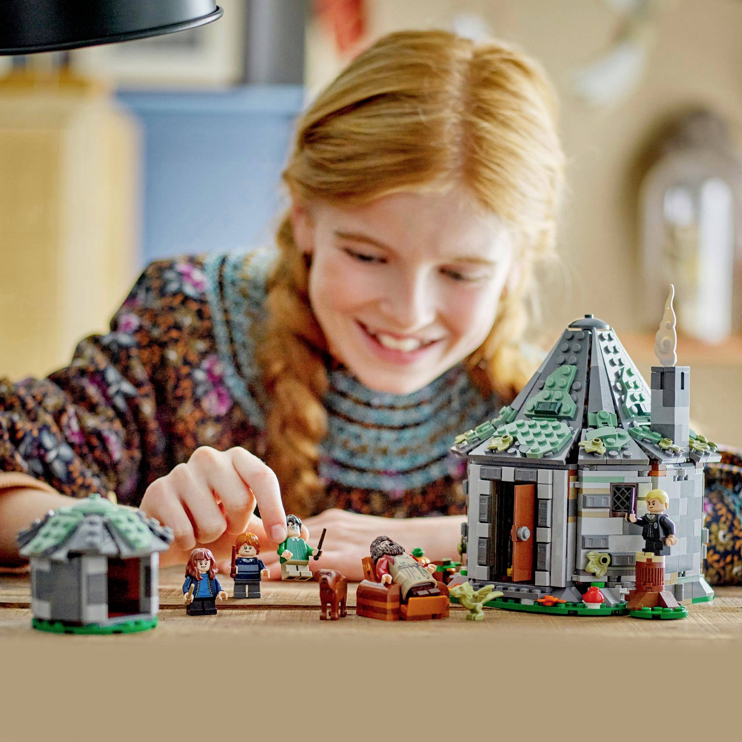 Ein Kind spielt mit einem Lego-Set in Form eines Hauses. Es lächelt und bewegt Figuren. Im Hintergrund ist ein unscharfer Raum zu sehen.
