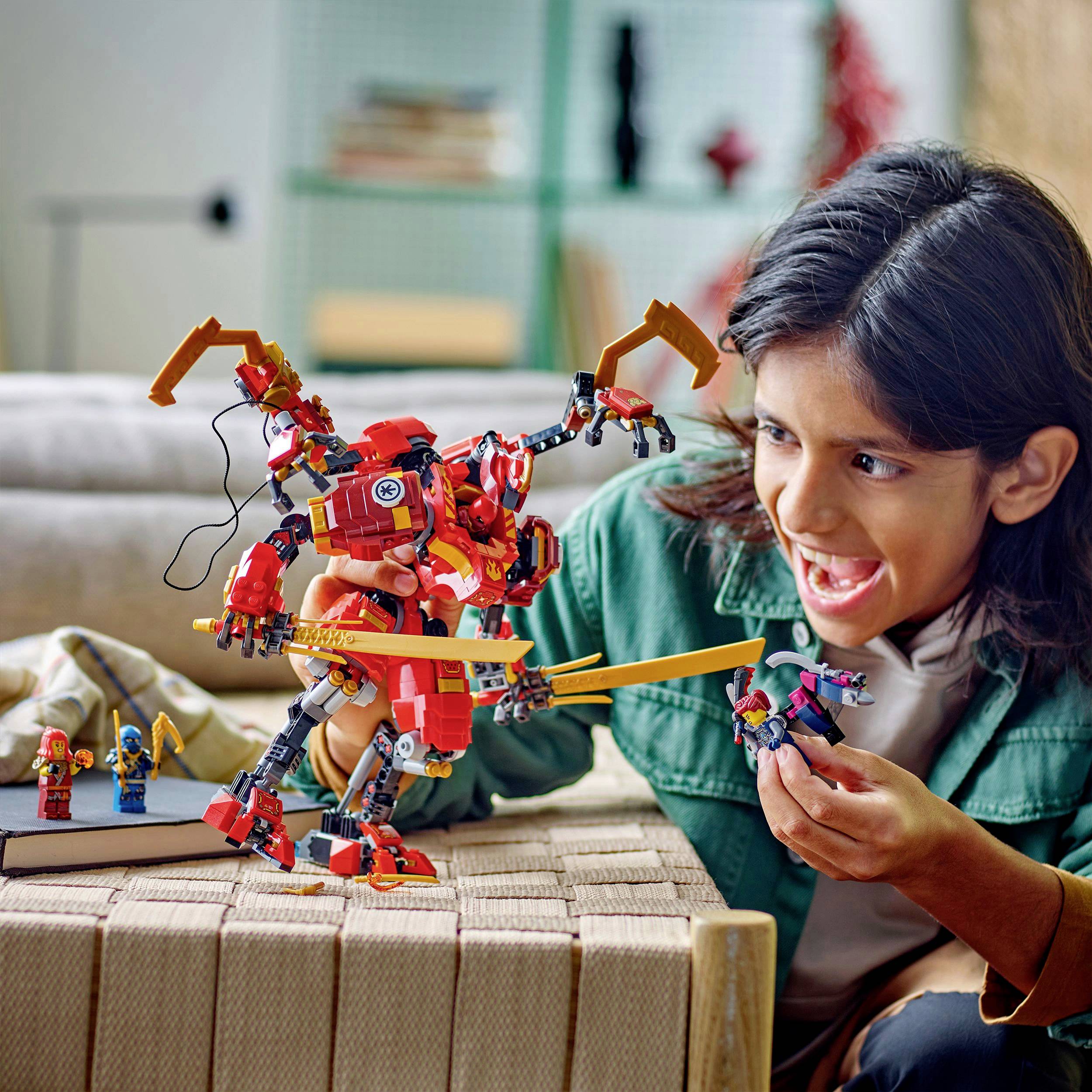 Ein Kind spielt begeistert mit einem großen, roten Roboter-Spielzeug auf dem Tisch. Im Hintergrund sind Bücher und ein Sofa sichtbar.