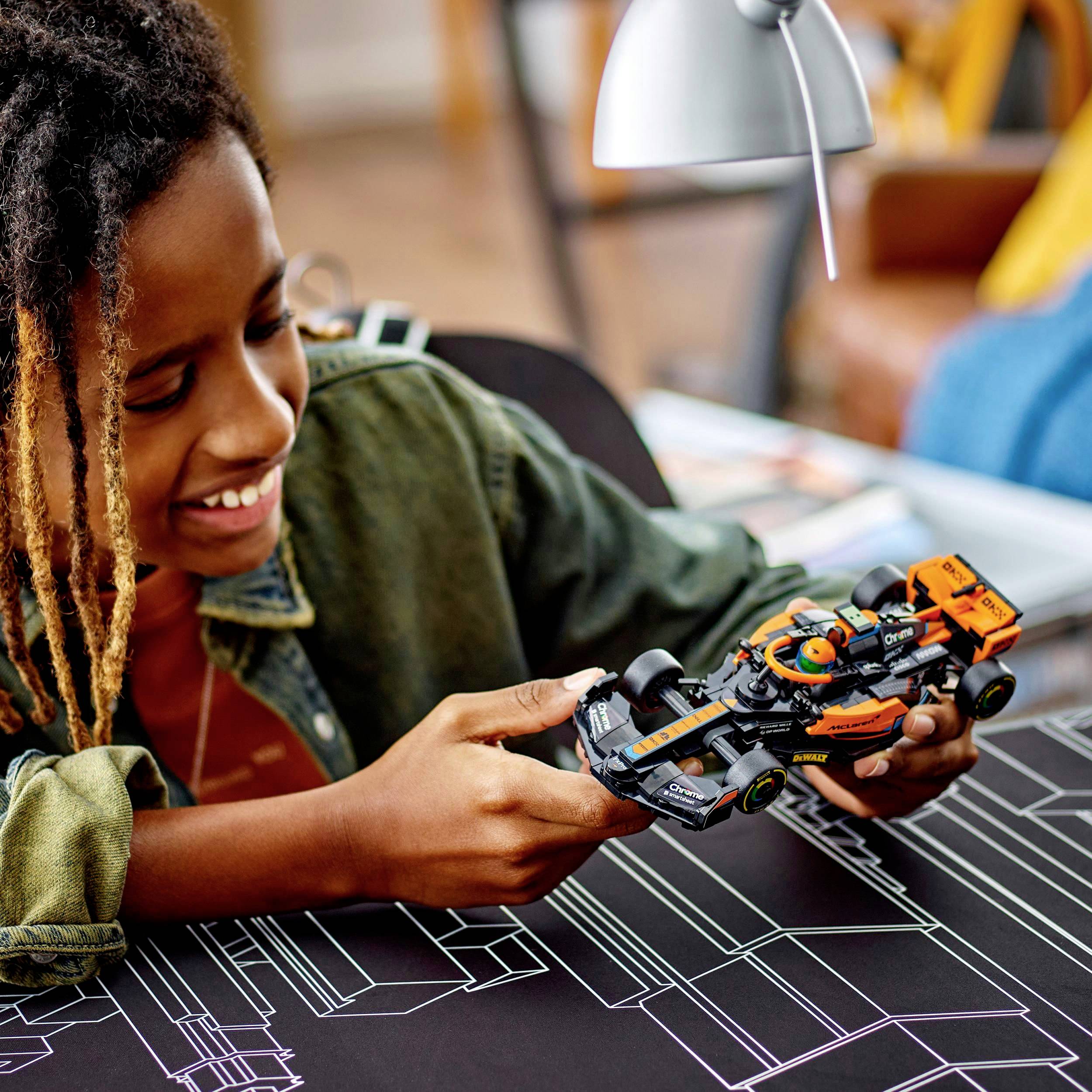 Ein Kind spielt lächelnd mit einem Modellauto auf einem Tisch, auf dem Gebäude-Skizzen gezeichnet sind. Im Hintergrund ist eine Lampe.