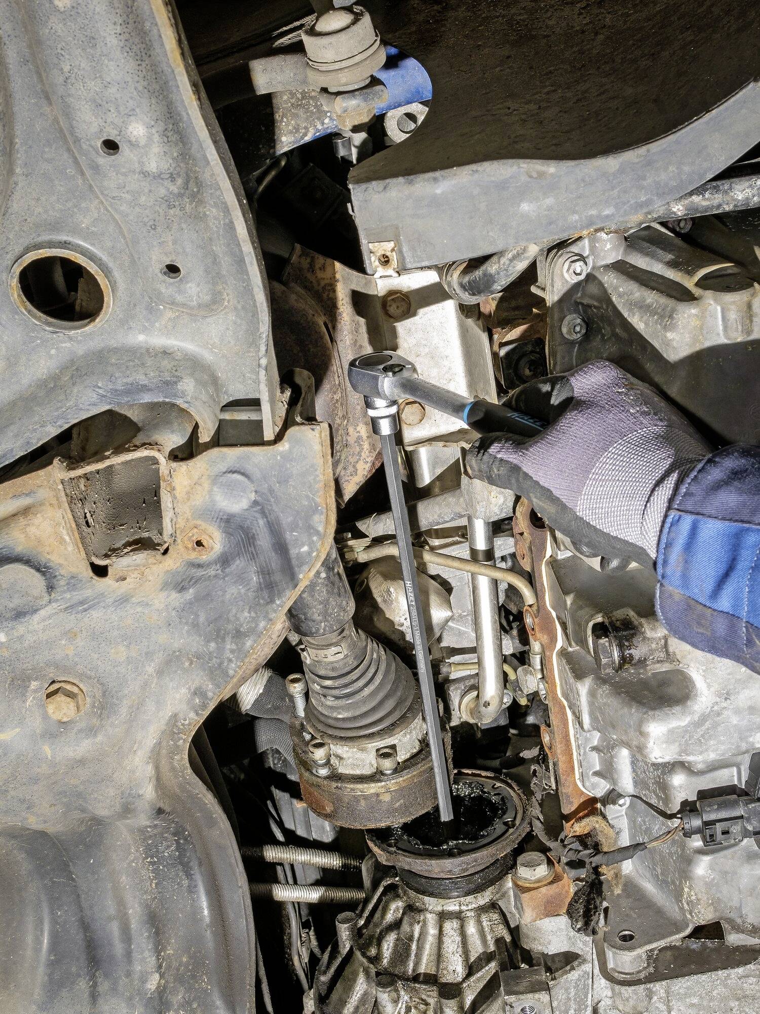 Eine Person repariert den Unterboden eines Autos, verwendet Werkzeug an der Achse, trägt Handschuhe und blau-schwarze Schutzkleidung.