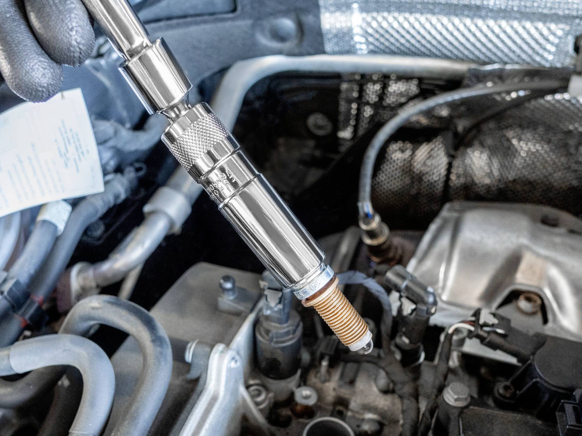 Zündkerze wird mit einem Werkzeug in den Motor eines Autos eingebaut. Etikett mit Text auf einer der Leitungen nebenan.