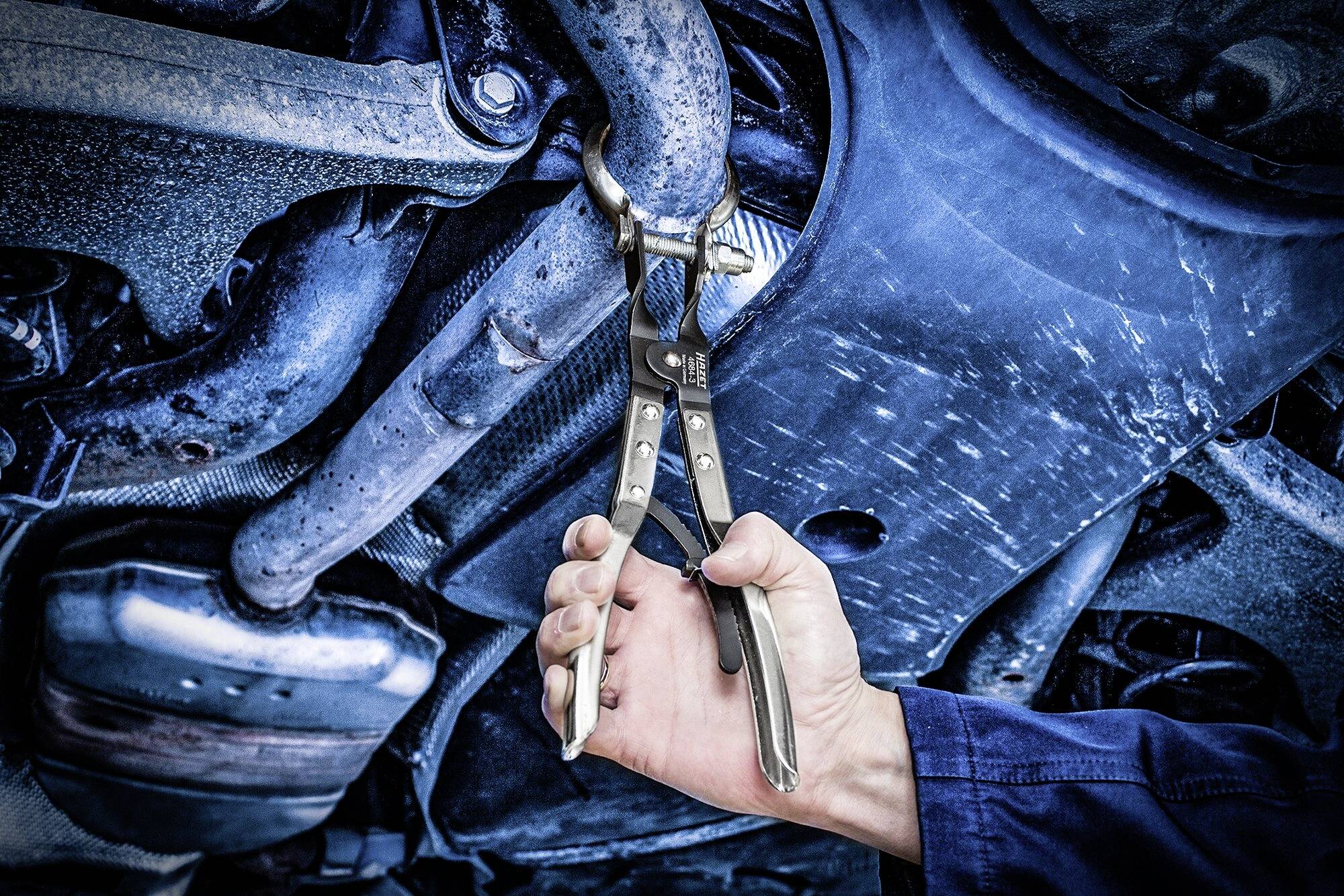 Eine Hand in Arbeitskleidung hält eine Zange, die am Auspuff eines Autos befestigt ist, während es repariert wird.