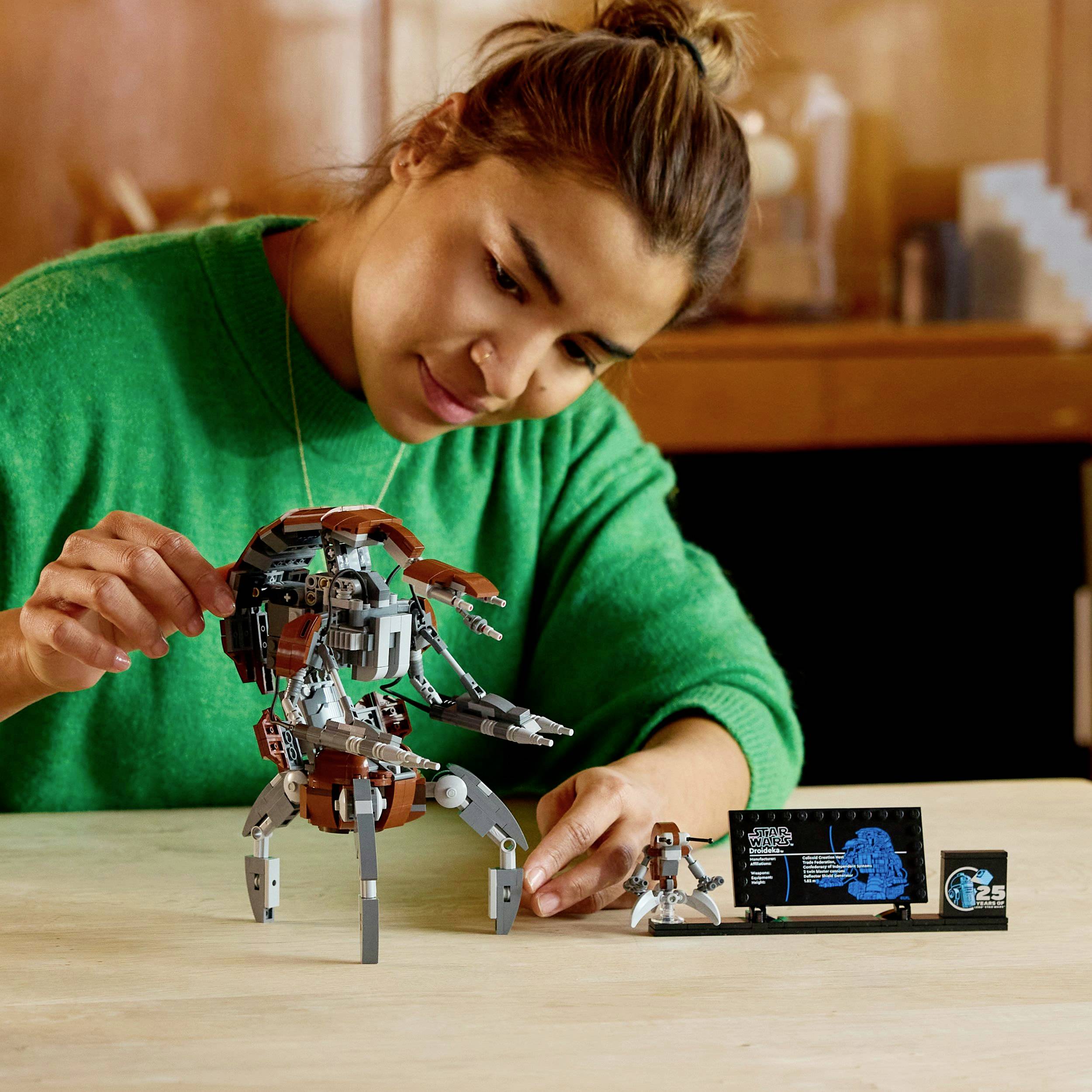 Eine Person baut ein futuristisches Roboter-Modell auf einem Tisch zusammen. Im Hintergrund sind Regale mit Wohnaccessoires zu sehen.