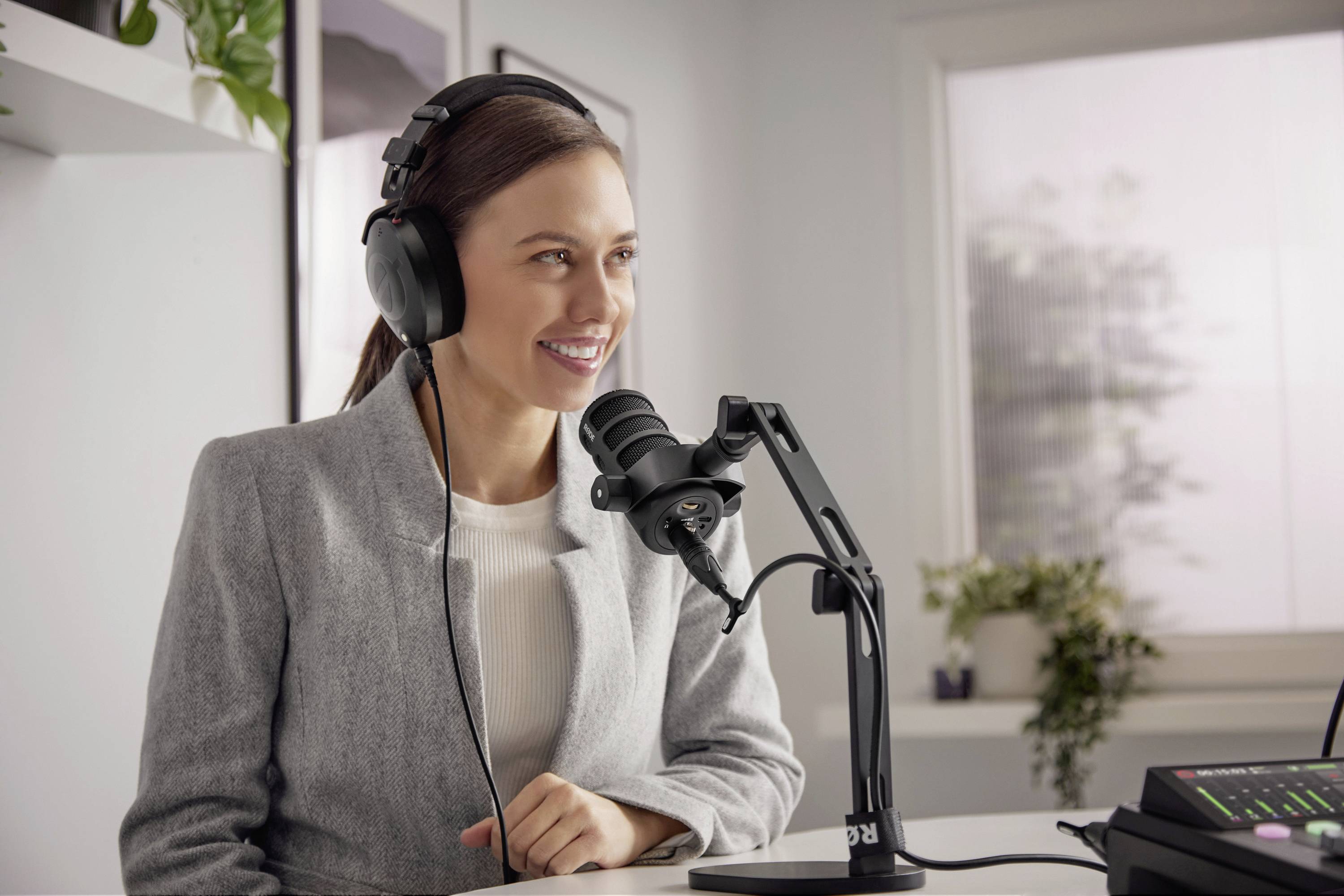 Eine lächelnde Frau in einem hellgrauen Blazer spricht in ein Mikrofon. Sie trägt Kopfhörer und sitzt in einem modernen Raum.