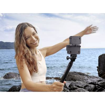 Eine Frau lächelt, während sie ein Selfie am Meer mit einem Smartphone auf einem Selfie-Stick macht. Im Hintergrund sind Felsen und Wasser.