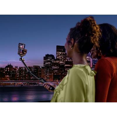 Zwei Personen stehen abends vor einer Stadtsilhouette und machen ein Selfie mit einem Selfiestick. Lichter der Stadt im Hintergrund.