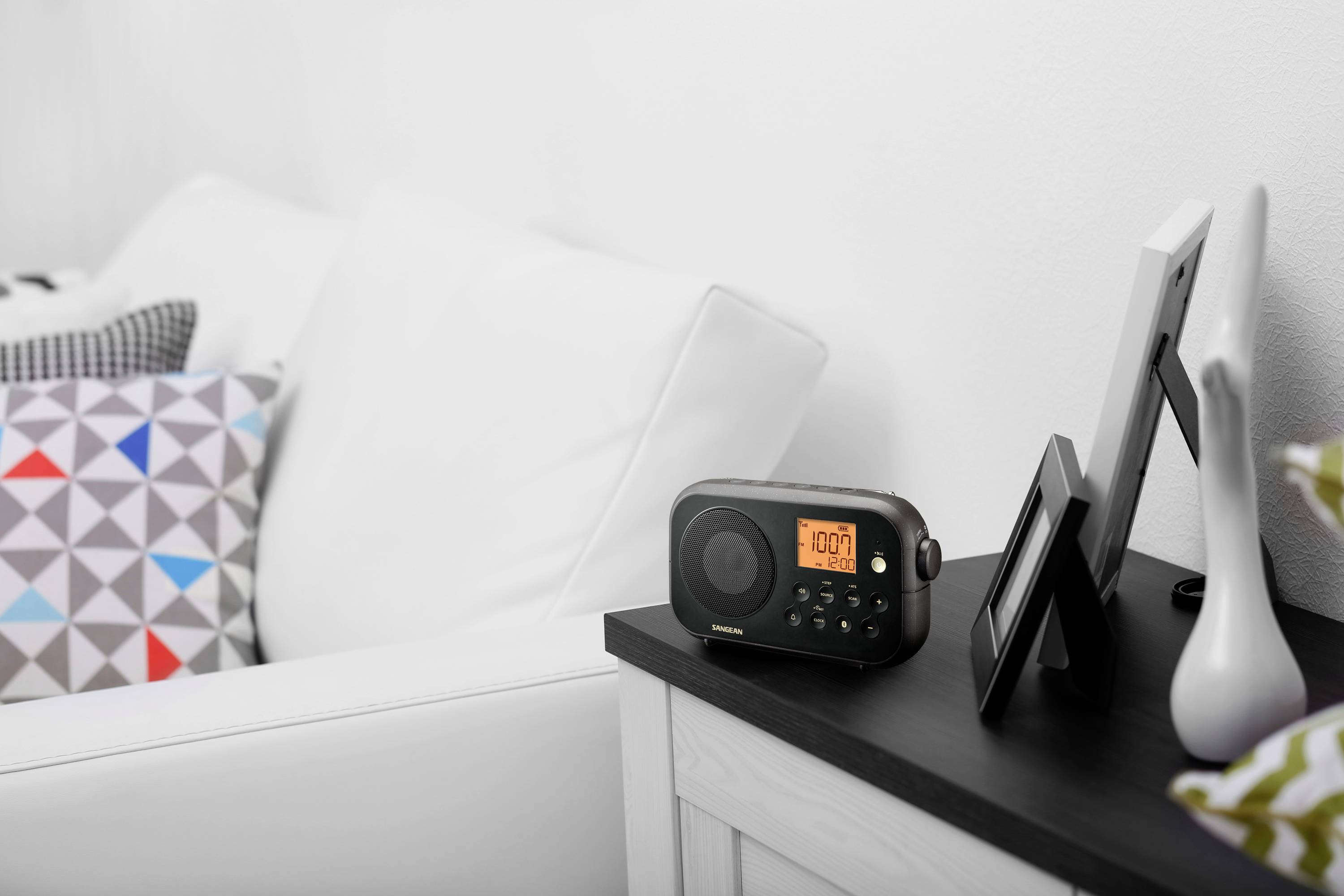 Radio auf einem Schrank, Uhrzeit 10:07 Uhr im Display. Neben dem Radio stehen Bilderrahmen. Ein Sofa mit Kissen ist im Hintergrund.