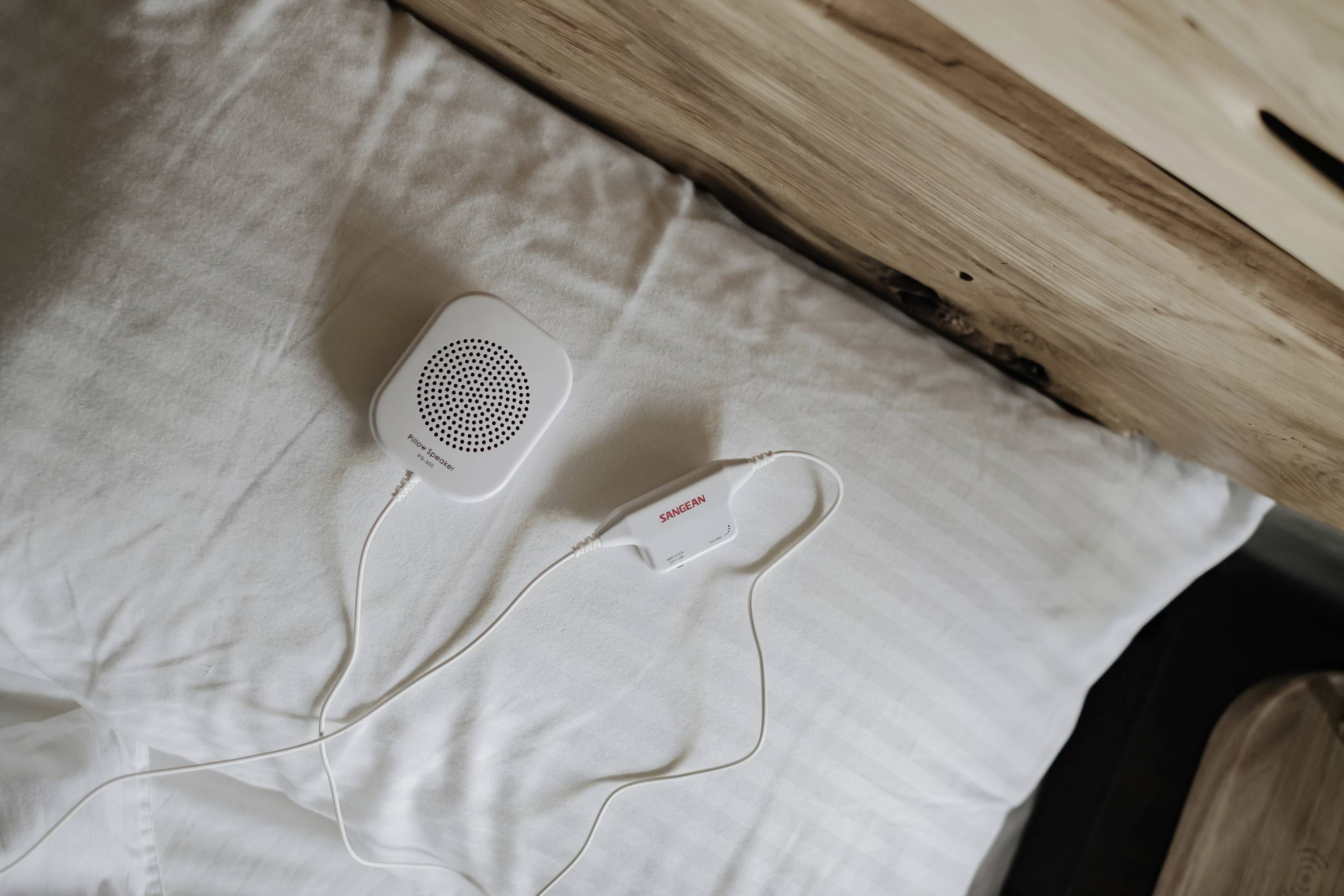 Ein weißes Babyphon liegt auf einem Bett mit einem weißen Kissen, Kabel verbunden. Holzoberfläche im Hintergrund ersichtlich.