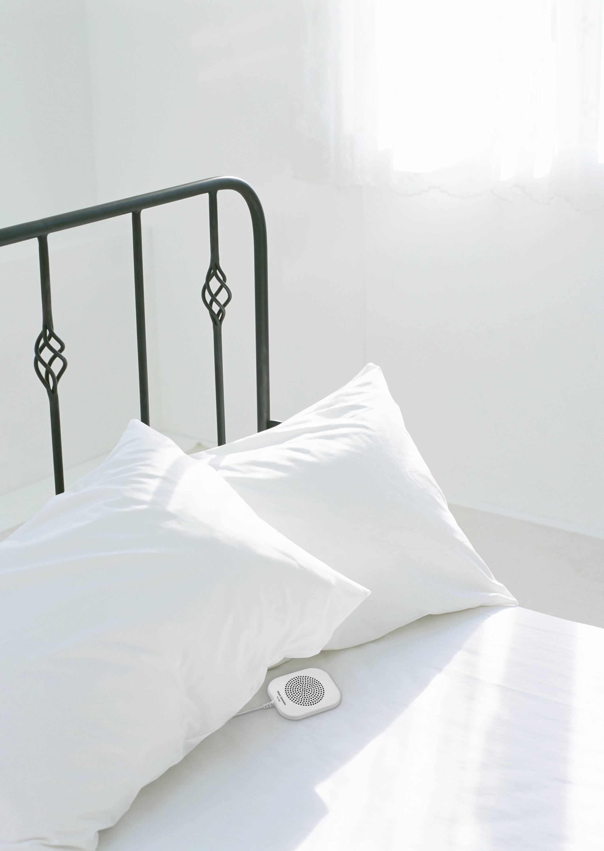 Ein weißes Bett mit zwei Kissen, durch Sonnenlicht von einem Fenster beleuchtet. Ein kleiner weißer Lautsprecher liegt auf dem Bett.