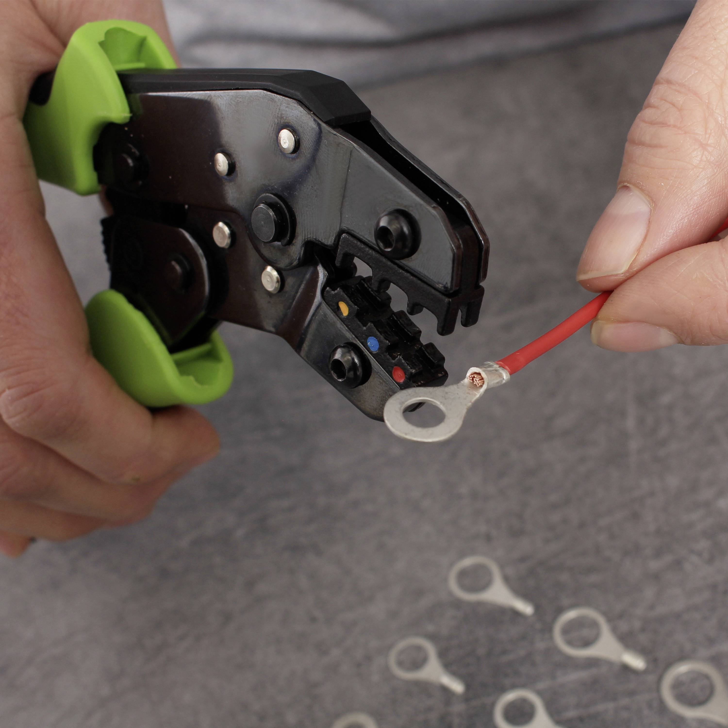 Eine Hand hält eine Crimpzange und crimpt ein rotes Kabel mit einem Ringkabelschuh. Im Hintergrund sind weitere Kabelschuhe zu sehen.