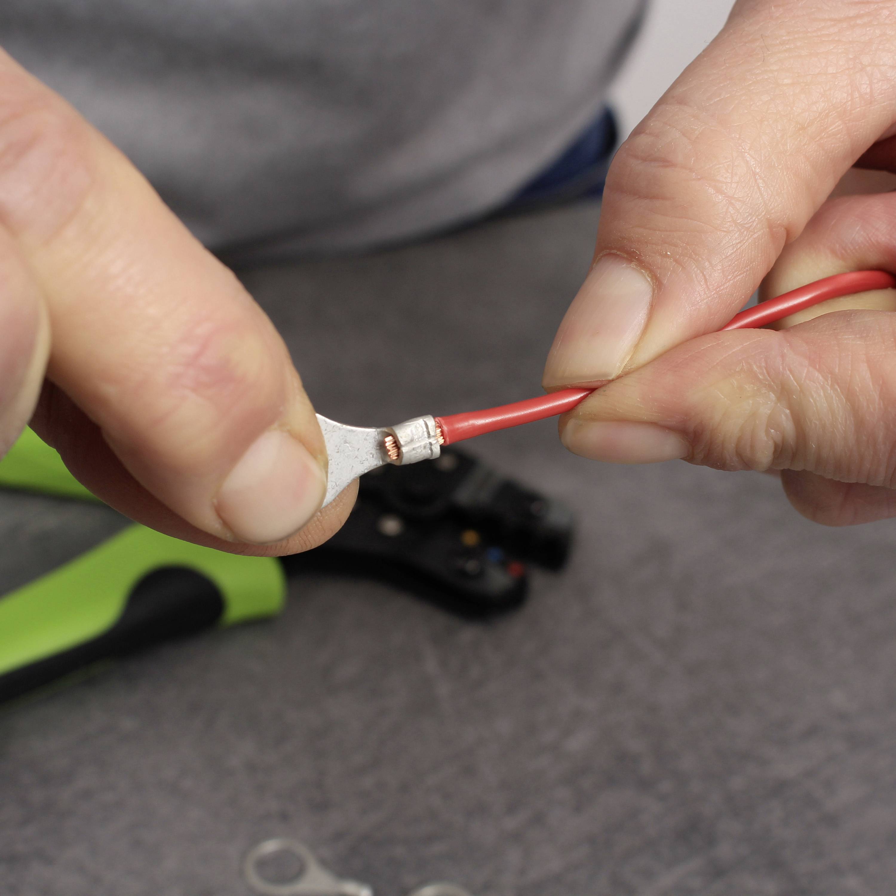 Eine Hand hält ein rotes Kabel mit einem am Ende befestigten isolierten Kabelschuh. Im Hintergrund liegt ein Werkzeug.