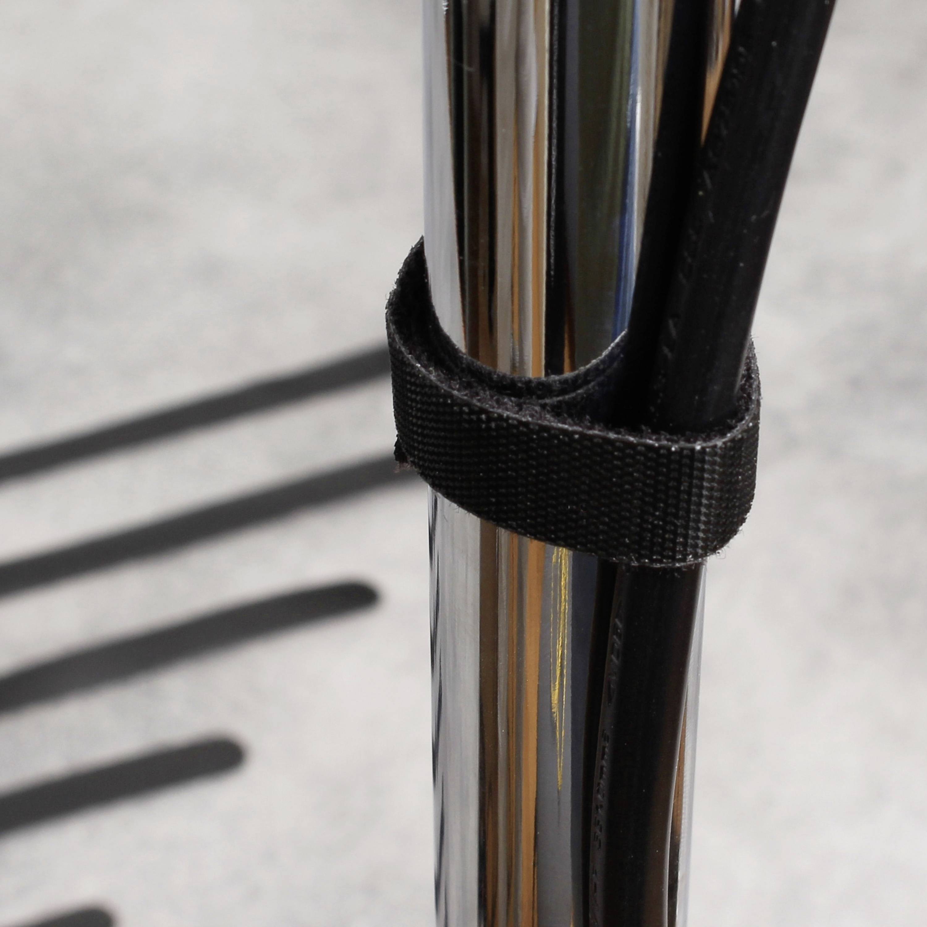 Ein schwarzes Kabel wird mit einem Klettverschluss um eine Metallstange befestigt. Im Hintergrund sind undeutliche Schatten zu sehen.