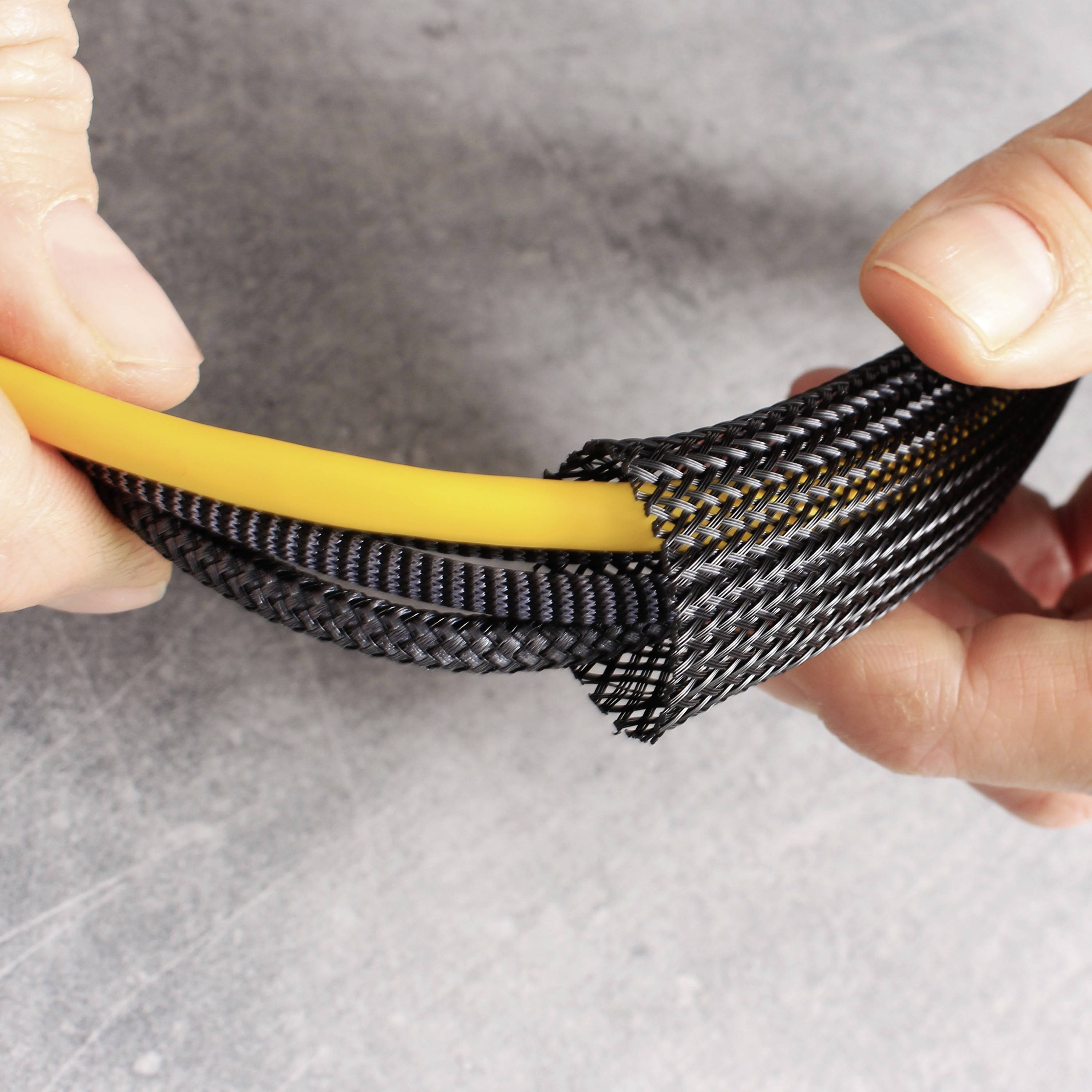 Eine Hand hält ein gelbes Kabel, das mit einem schwarzen, geflochtenen Kabelschutz umwickelt ist. Hintergrund ist grau.
