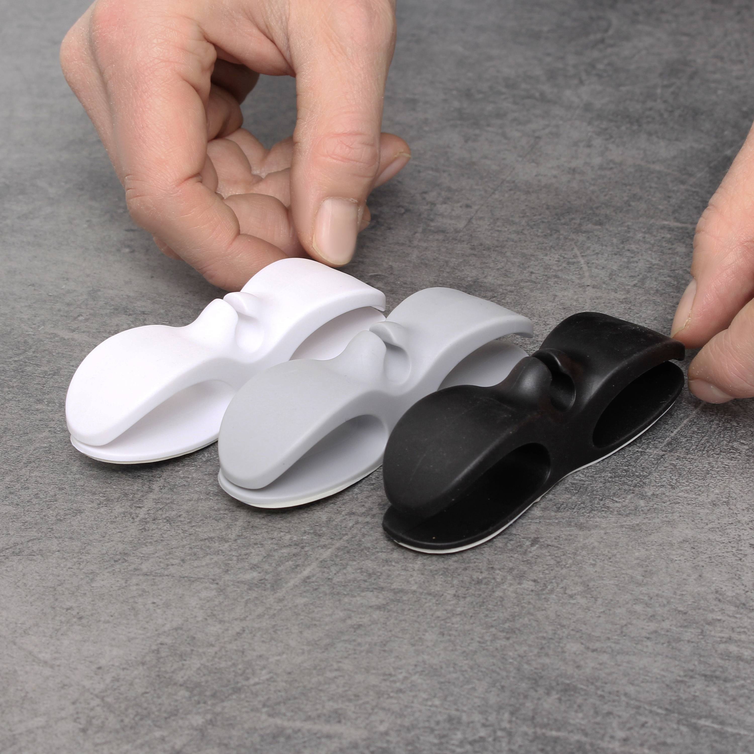 Drei Kabelhalter in Weiß, Grau und Schwarz auf grauer Oberfläche. Zwei Hände ordnen die Halter.